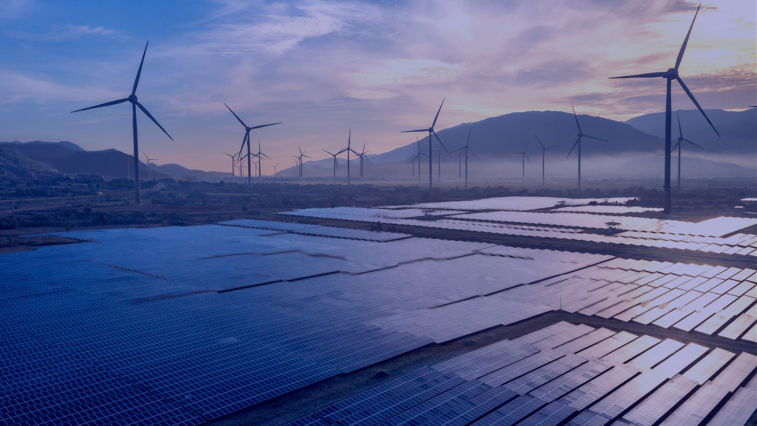 A landscape view of a solar farm and wind turbines set against mountains at sunrise or sunset, with mist over the ground.