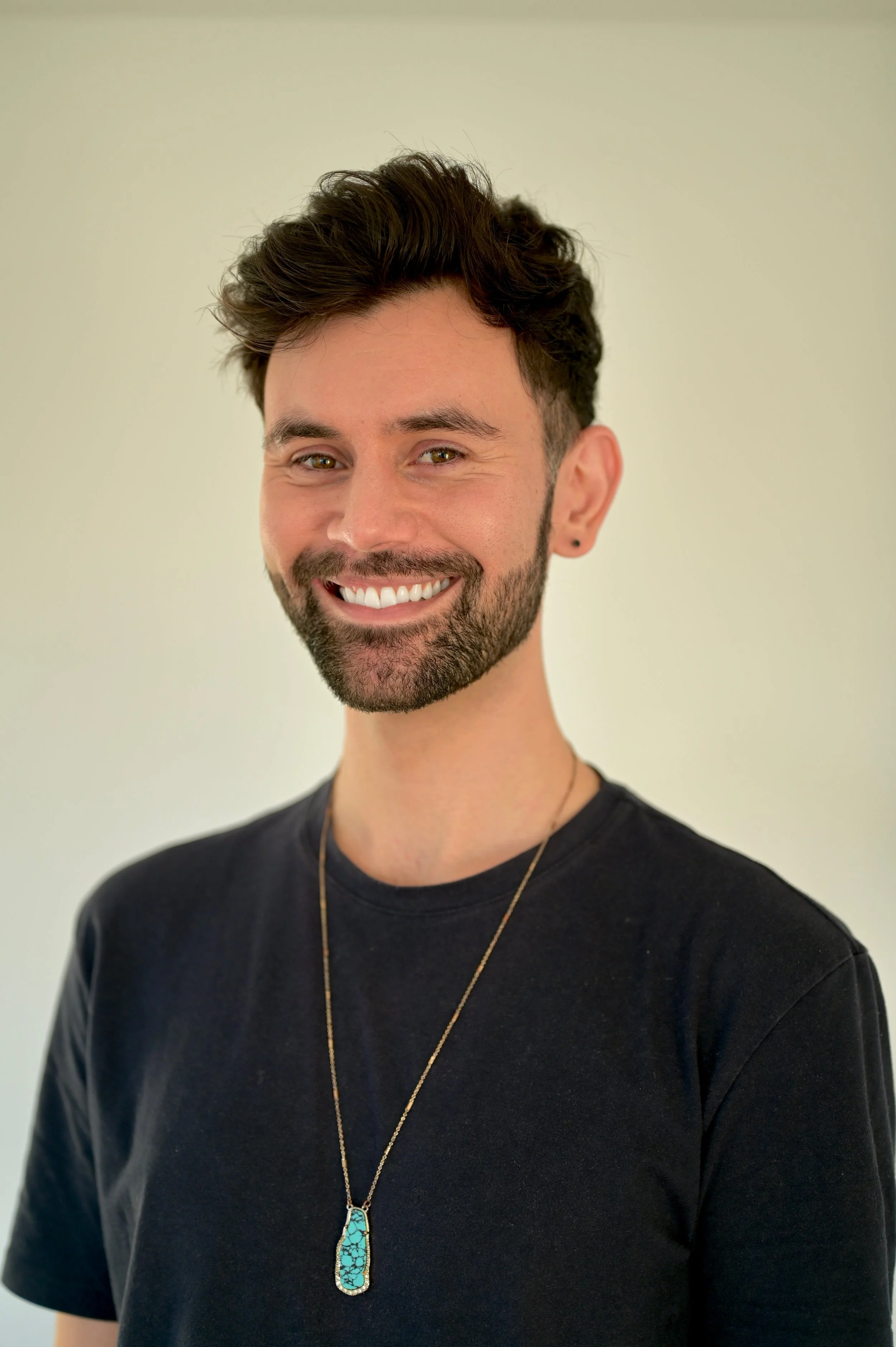 A smiling man with dark hair, a beard, wearing a black T-shirt, a necklace with a turquoise pendant, and a small earring in his left ear, standing against a light-colored plain background.