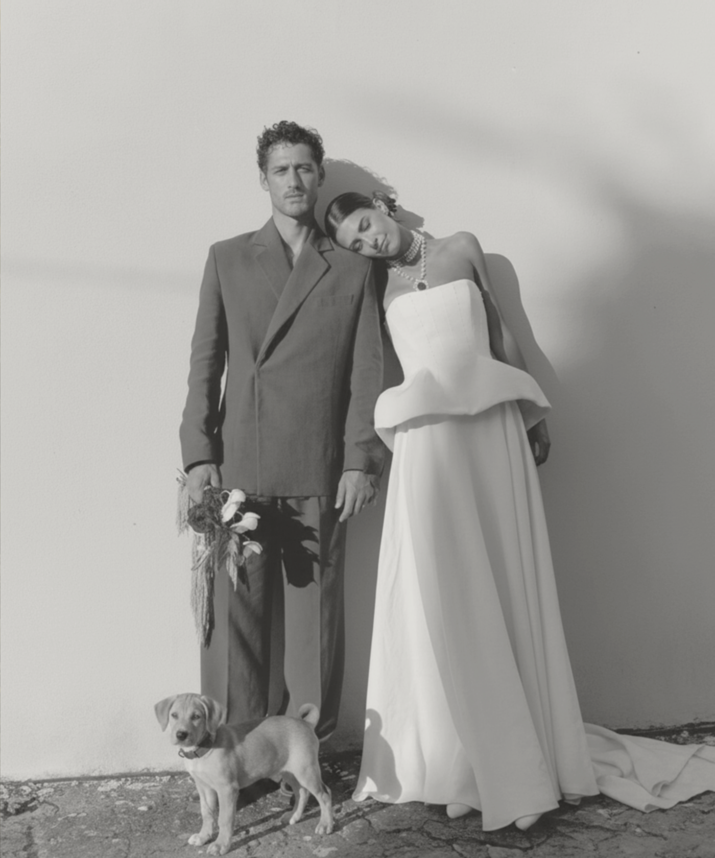 A black and white photo of a man and woman dressed in formal wedding attire standing next to each other. The woman is resting her head on the man's shoulder. The man is holding a small bouquet of flowers and the woman is wearing jewelry and a strapless gown. A small puppy stands in front of them, looking at the camera. The background is plain and minimalistic.