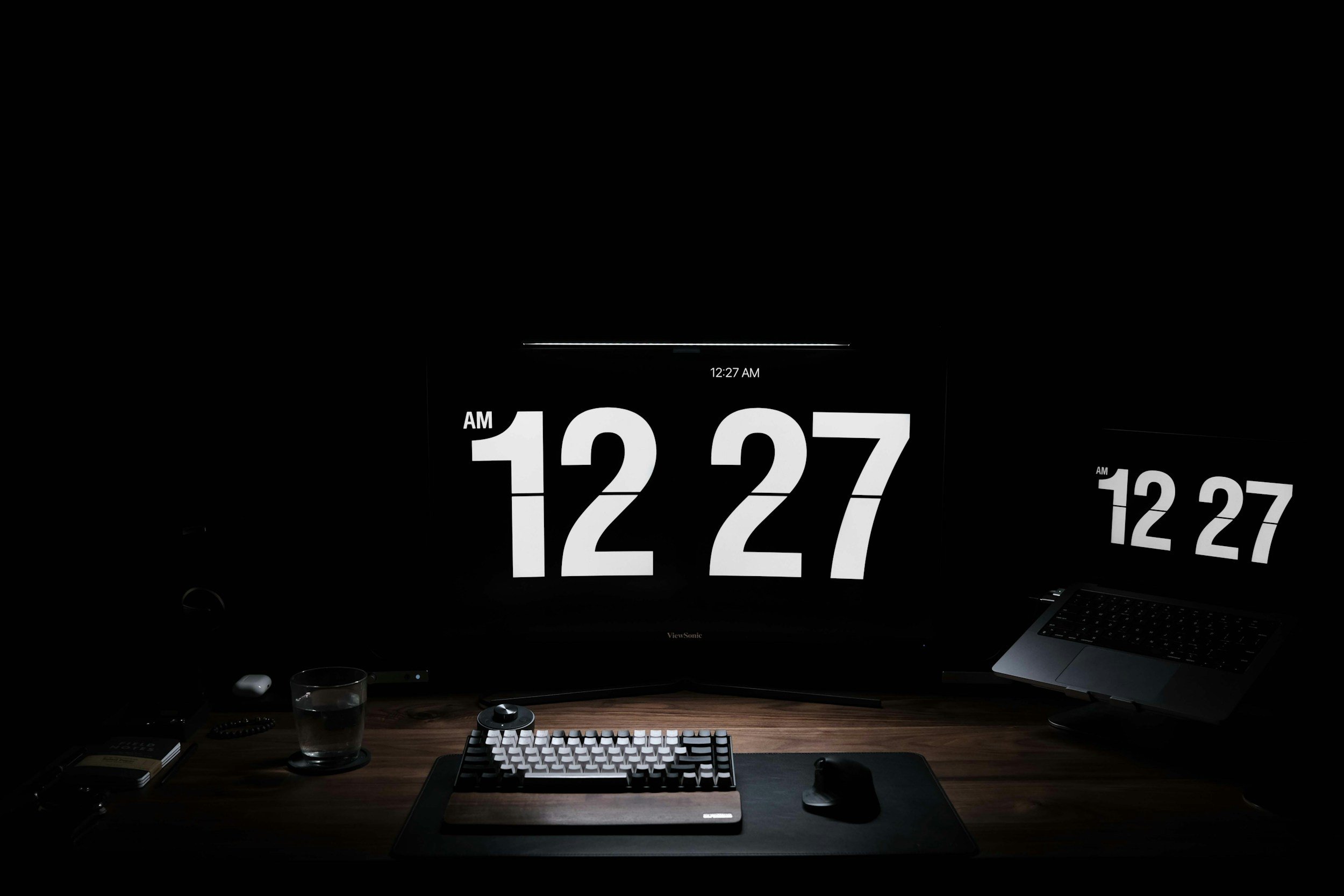 A dark workspace with a large central monitor displaying the time 12:27 AM in bold white numbers, reflected on a secondary monitor to the right. The desk has a mechanical keyboard, a mouse on a mouse pad, a glass of water, and a few other small items. A laptop is open on the right side.
