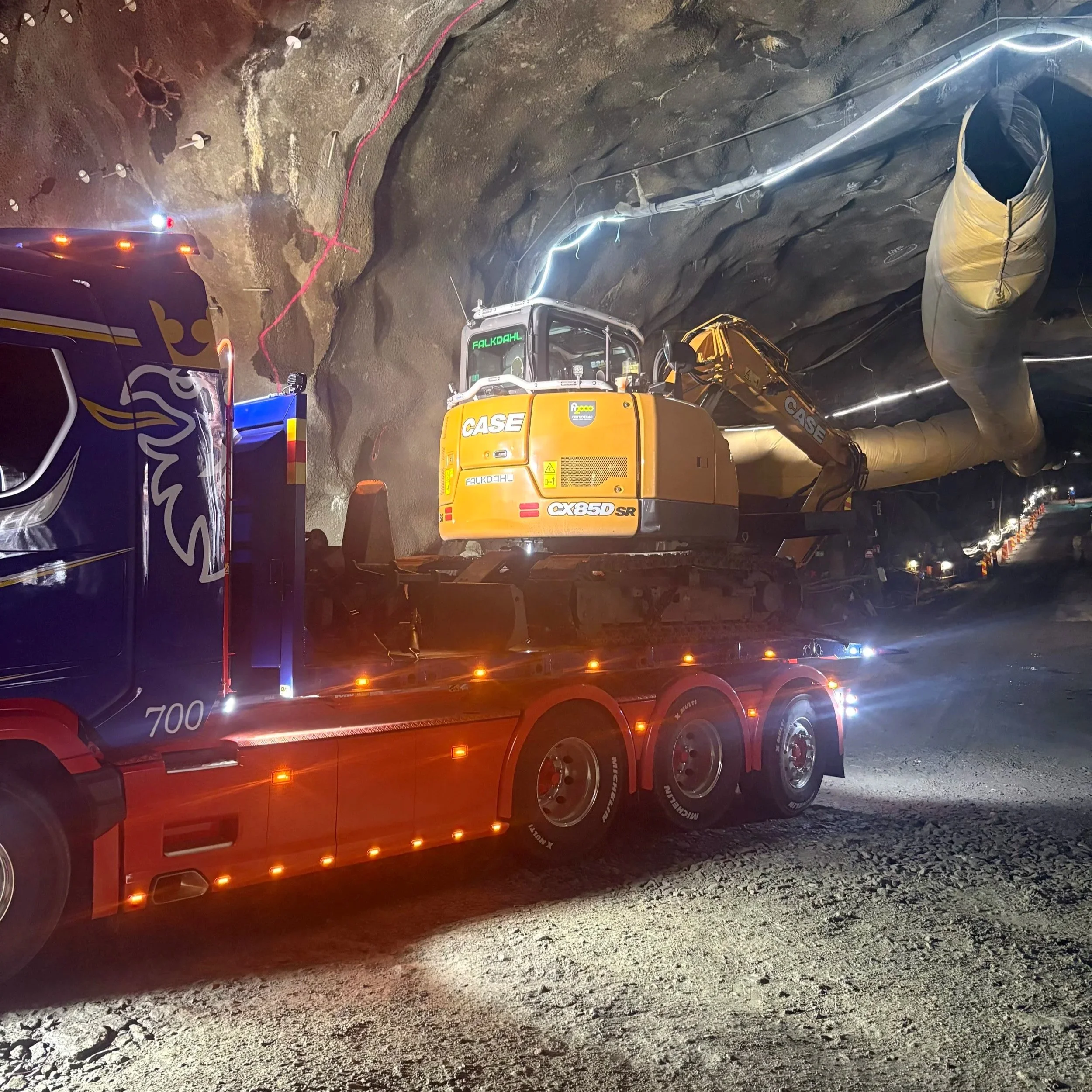 En grävmaskin som arbetar i en grotta eller tunnel, med olika kablar och utrustning synlig. Belysning avger ett blått sken i den mörka miljön.