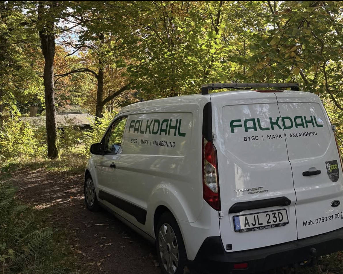En vit arbetsbil med företagsnamnet Falkdahl BYGG, MARK, ANLÄGGNING skrivet på sida och bak i en skogsmiljö.