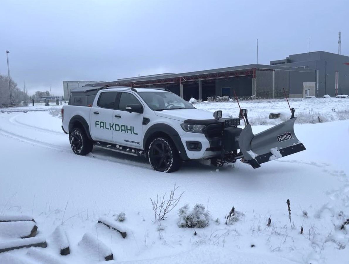 Vit fordon med snöskyffel på vagnsidan, texten 'Falkdahl' och 'Hilltip' på snöskoveln, parkerad på snöigt område med industribyggnader i bakgrunden.