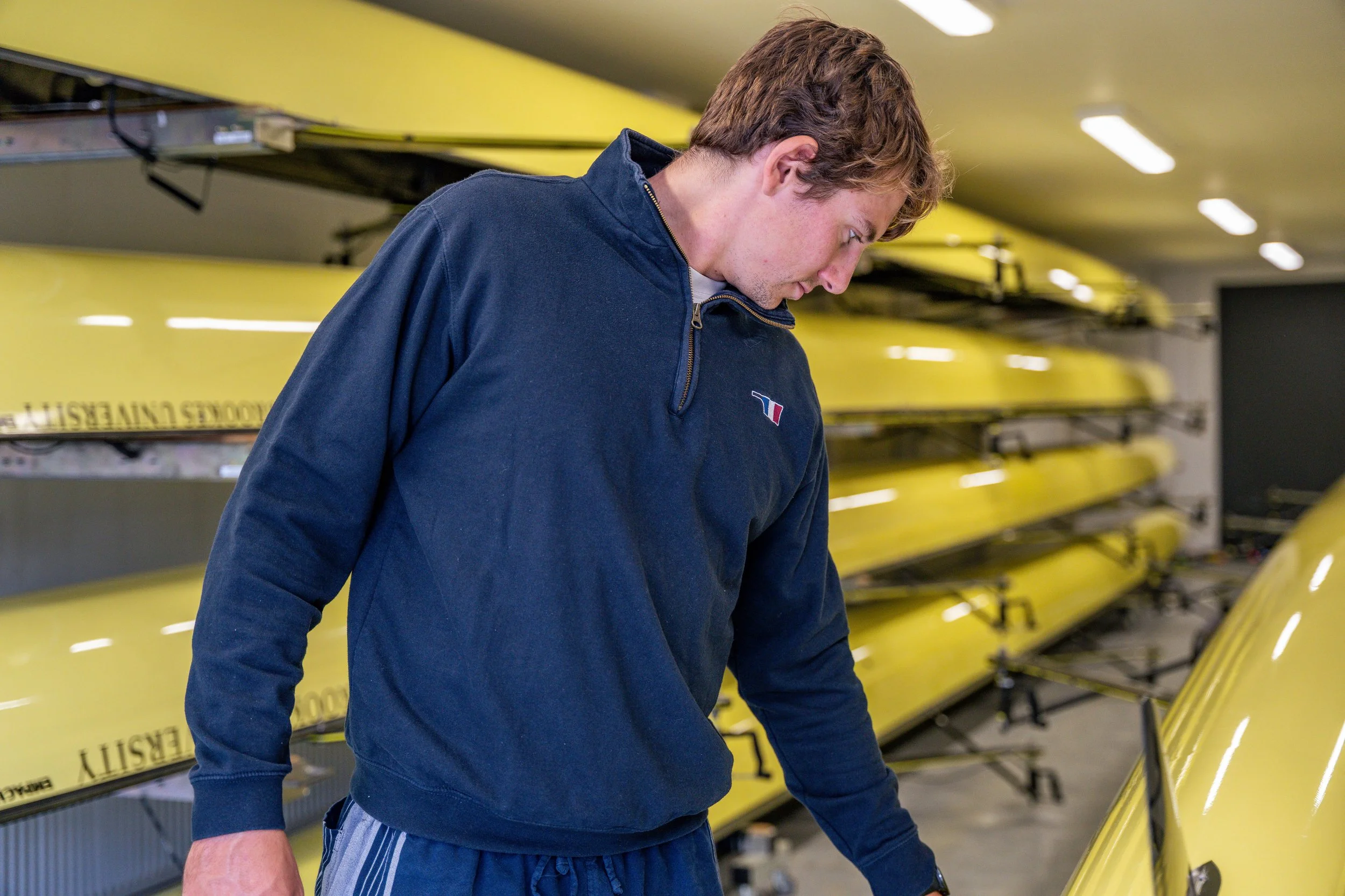 A young man with light brown hair wearing a navy blue pullover looking down, standing in front of yellow rowing shells in a storage area.
