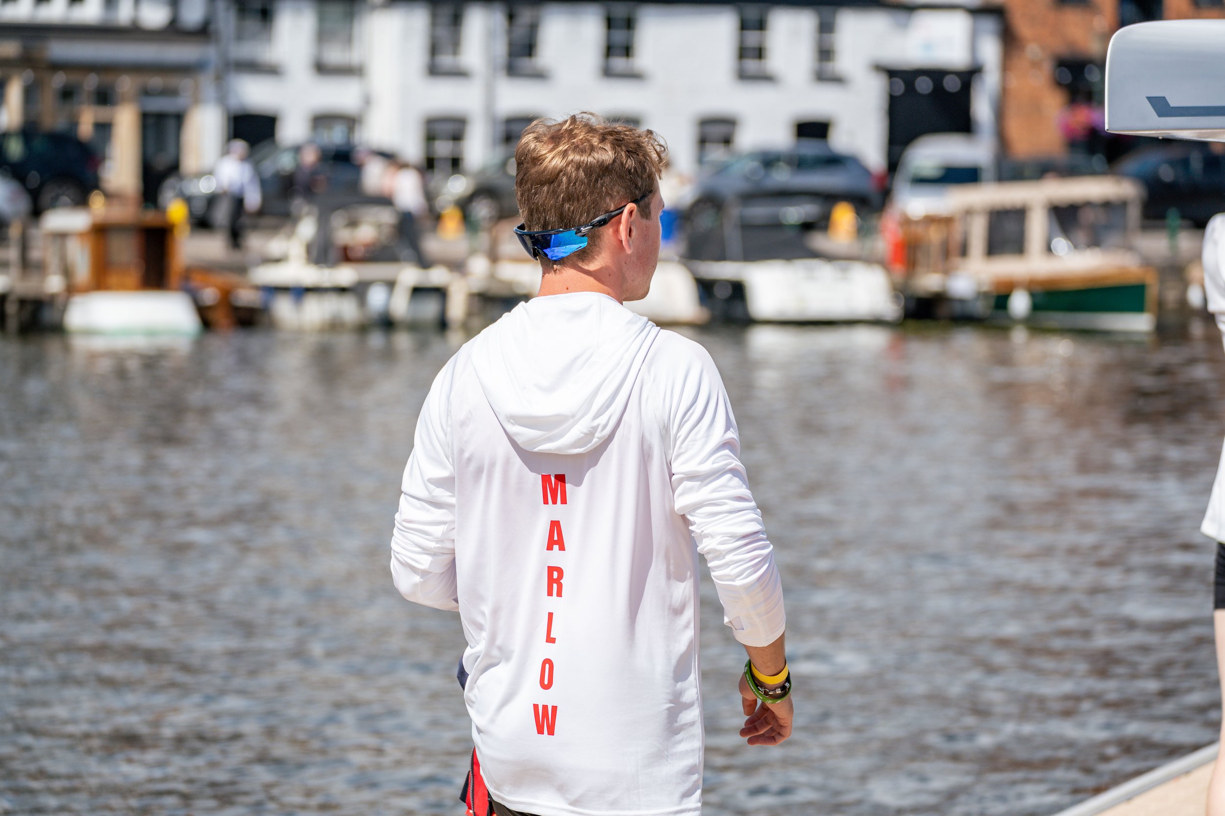 A young man with brown hair wearing sunglasses and a white hoodie with red text on the back, walking along a waterfront with boats and buildings in the background.