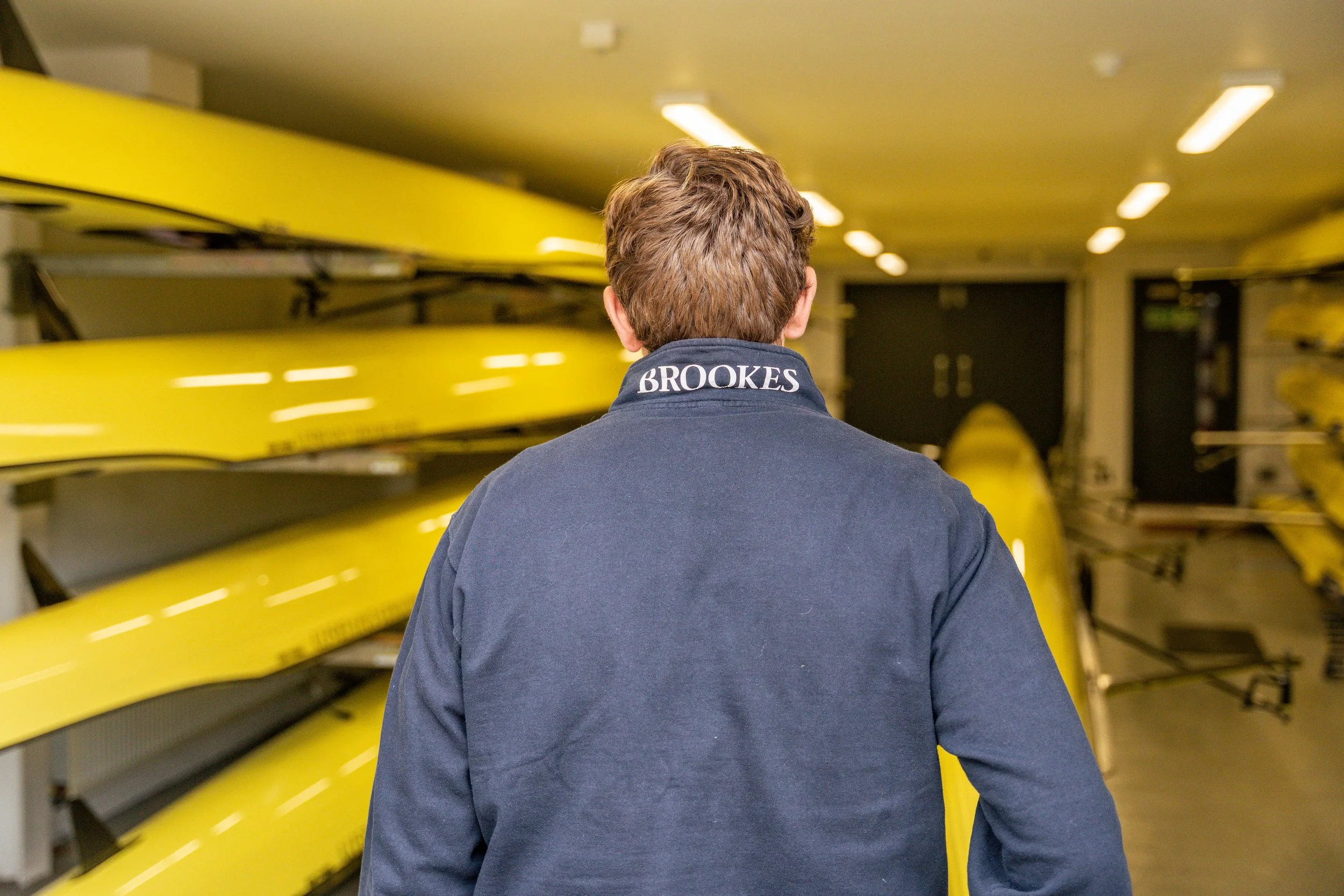 A person with brown hair wearing a navy jacket with the name 'Brookes' embroidered on the collar, standing in front of yellow rowing shells stored on racks in a rowing club or sports facility.