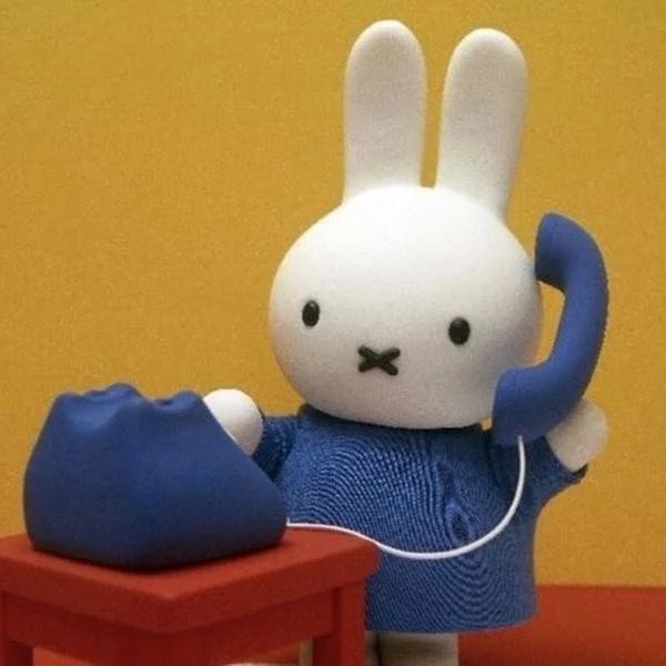 A plush bunny character holding a blue telephone receiver with a cord, standing beside a small red table with a blue telephone on it, against a yellow background.