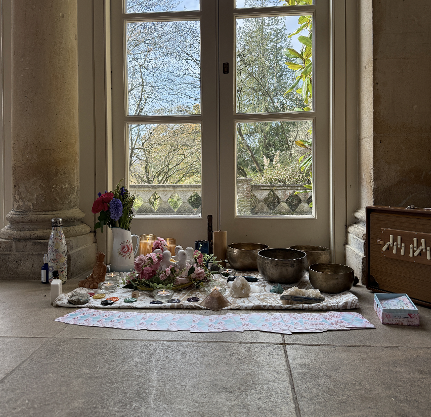 A collection of healing crystals, singing bowls, flowers, and spiritual items arranged on a cloth in front of a window with trees outside.