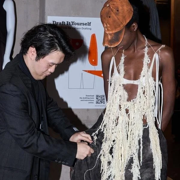 A man and a woman look at a dress made of shredded fabric or strings, with a backdrop featuring a poster about drafting or pattern creation.