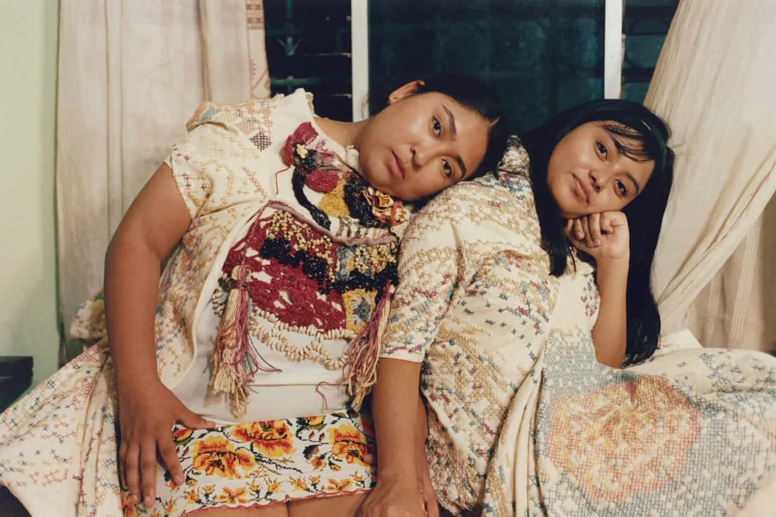 Two women lie on a bed with patterned bedding. One woman has her head resting on the other's shoulder, and both are looking at the camera. The woman on the left has her eye makeup done and is wearing embroidered clothing, while the woman on the right has her head propped on her hand and is smiling slightly.