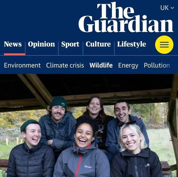 Group of six smiling young people outdoors under a wooden shelter, some wearing jackets and beanies, with trees in the background.