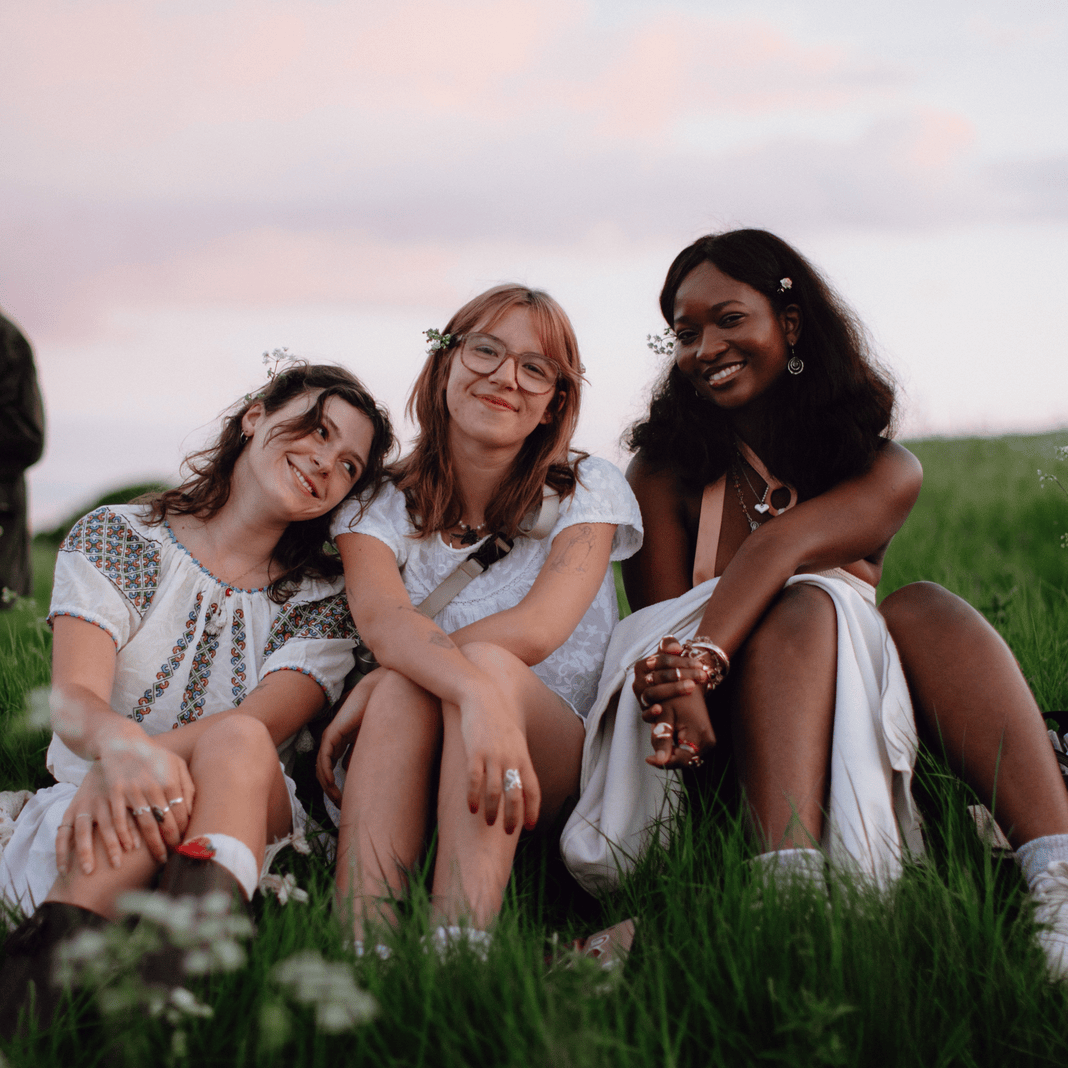 Three women sitting on grass in a field during sunset, smiling and enjoying each other's company.