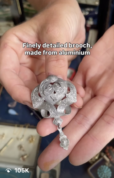 Person holding a detailed brooch made from aluminum, with the text 'Finely detailed brooch, made from aluminum' overlaid.