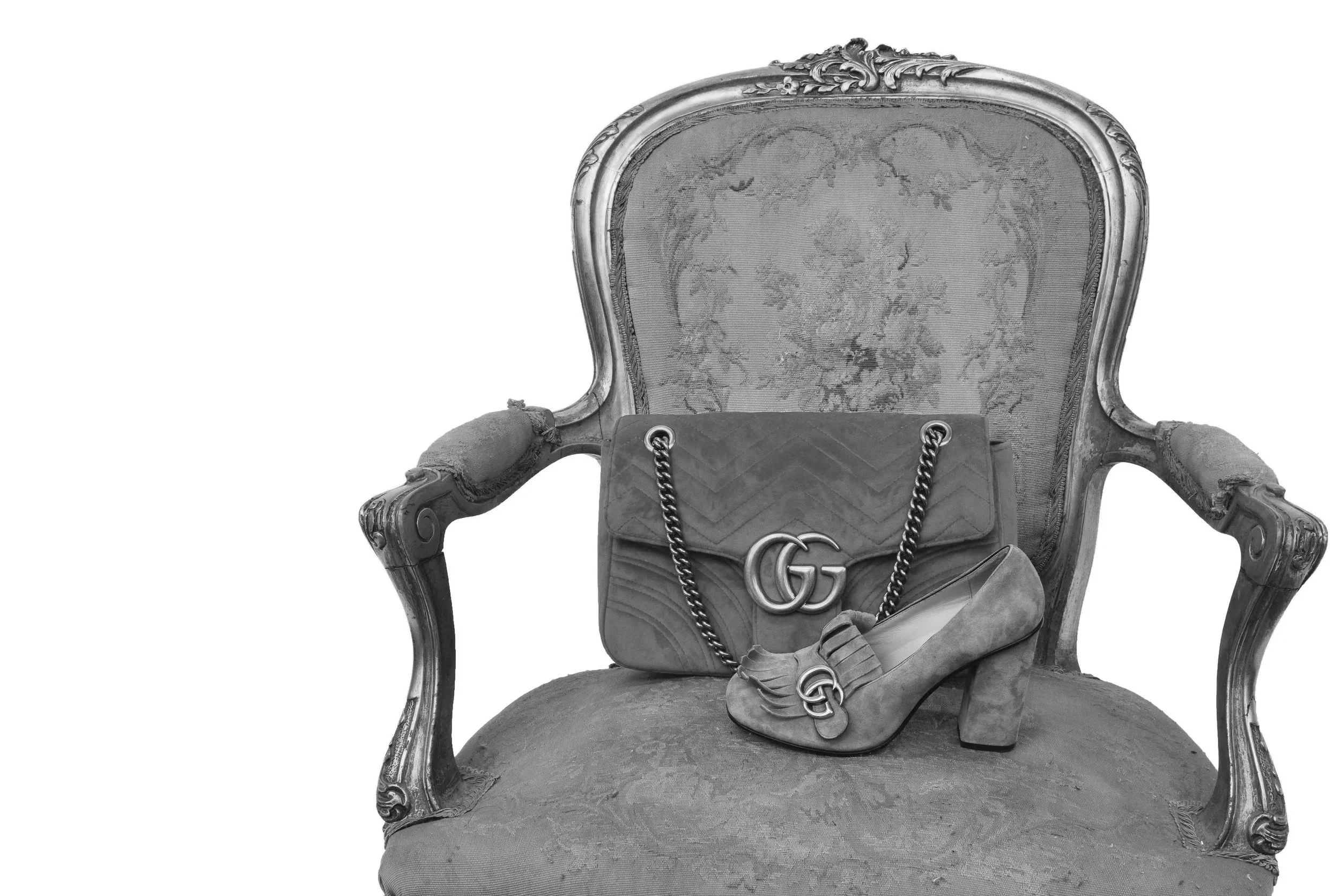An ornate vintage armchair with elaborate carved wooden frame and upholstery, featuring a Gucci handbag with chain straps placed on the seat.