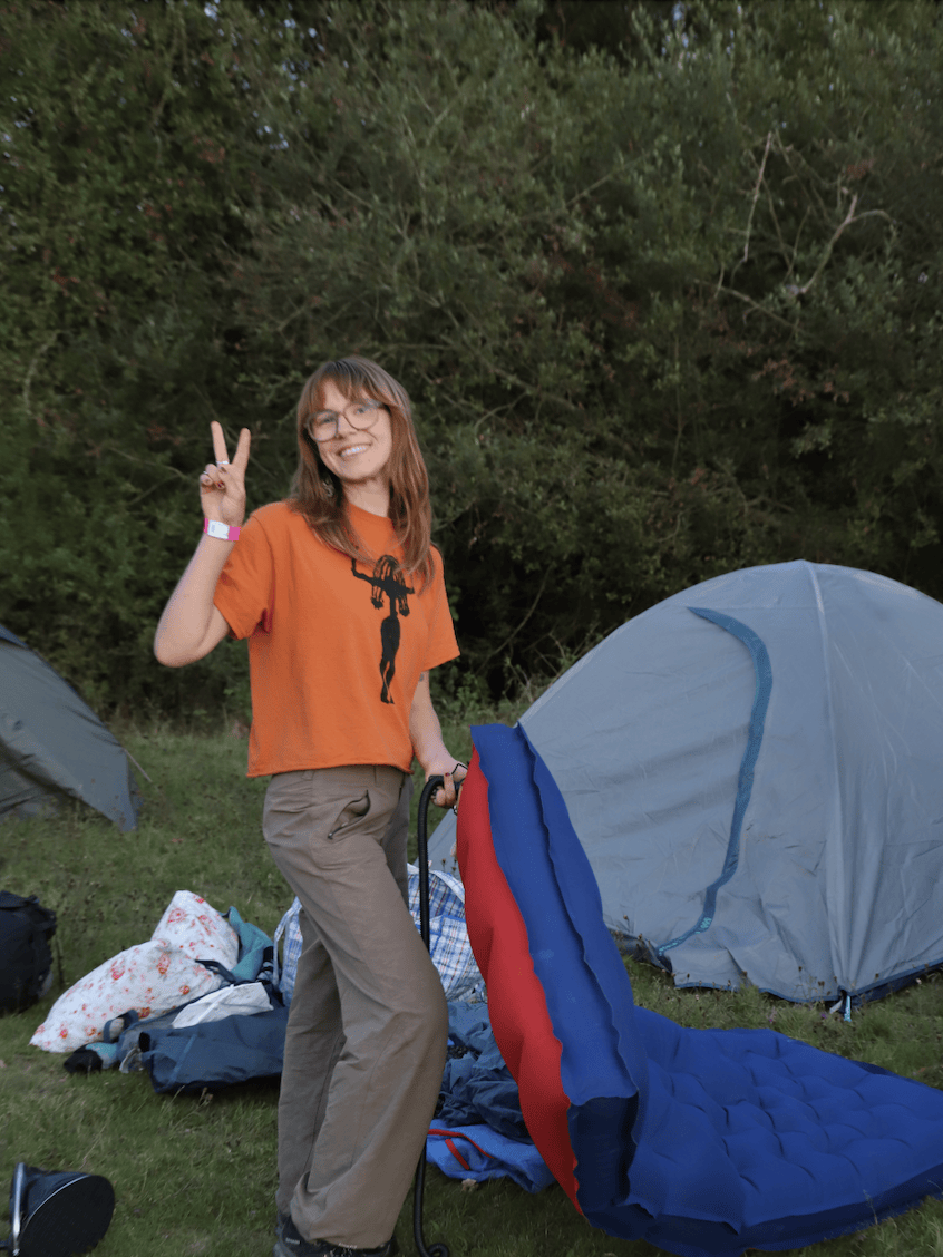 A woman with glasses smiling and making a peace sign outdoors near camping tents with an inflatable sleeping pad.