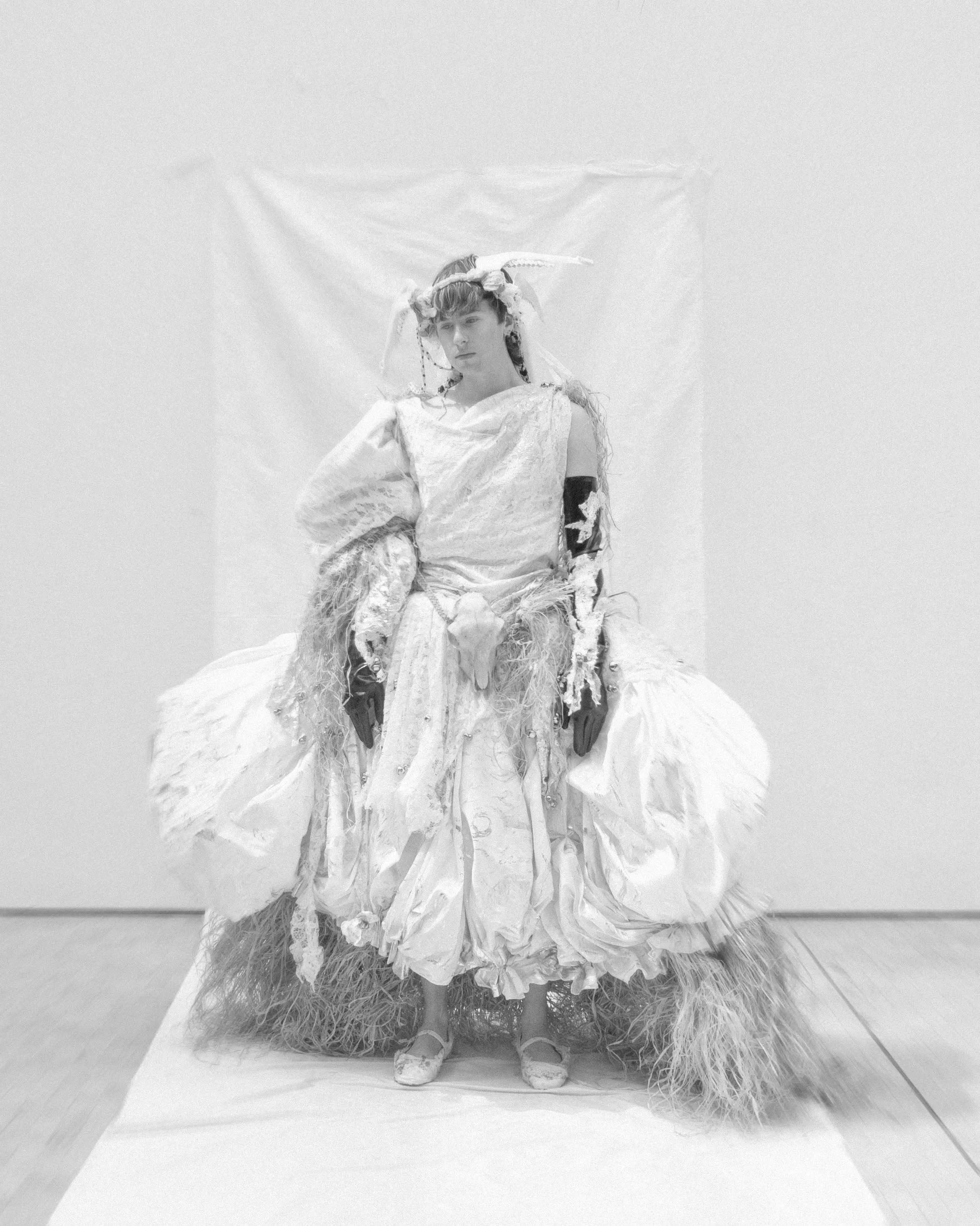 A woman dressed in elaborate, textured white garments stands against a white background, wearing a crown with an intricate design and long ribbons. Her outfit features layered fabrics, fringe, and detailed textures, with her hands visible but partially obscured by the clothing. The overall scene has a monochromatic, artistic aesthetic.
