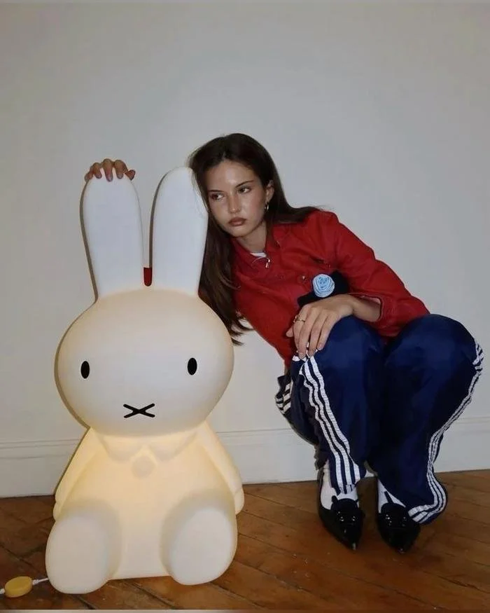 A young woman in a red jacket and blue sports pants is squatting next to a large illuminated bunny-shaped lamp, inside a room with wooden floors and plain white walls.