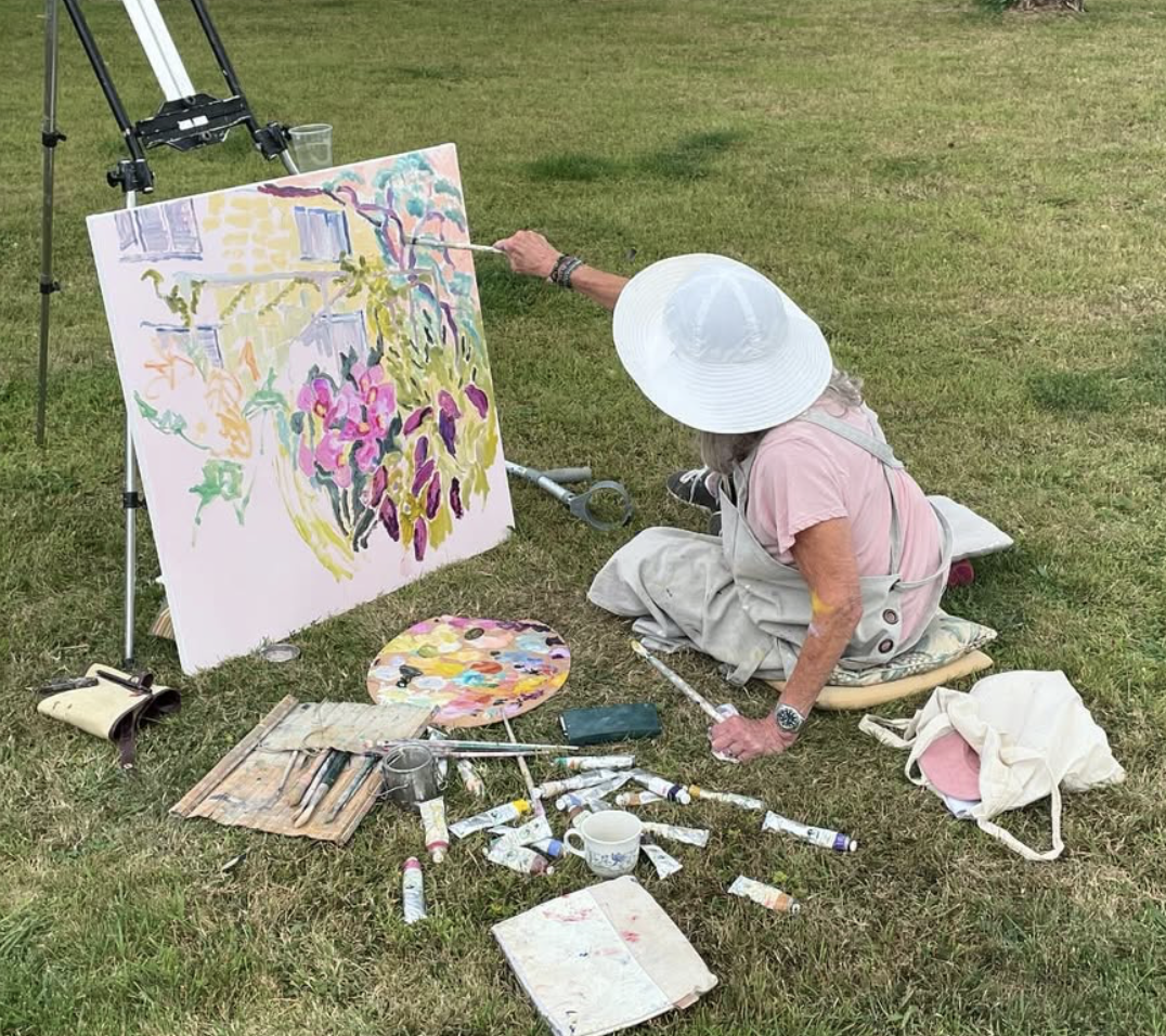 An elderly woman painting an outdoor landscape on a large canvas with various art supplies scattered around on the grass.