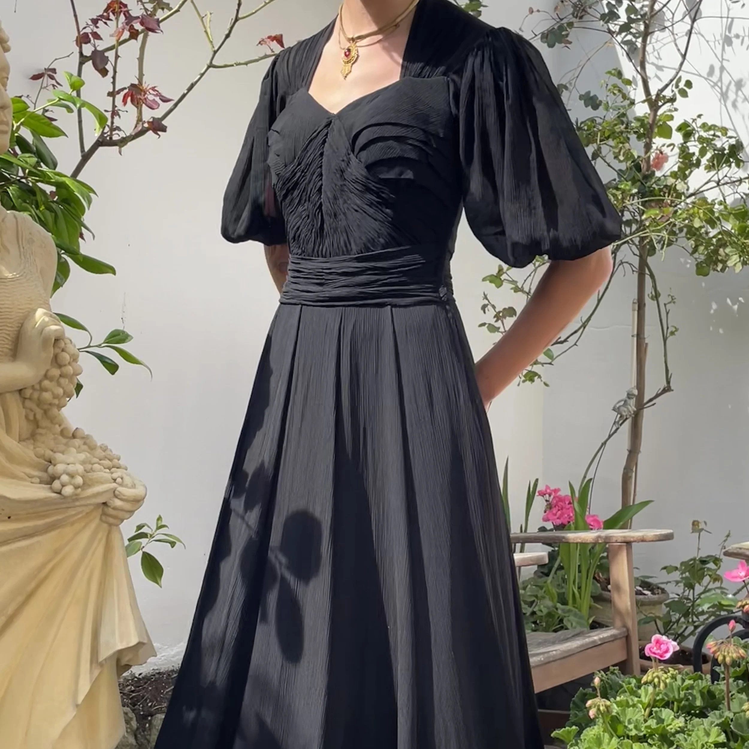 A person wearing a dark pleated dress and a gold necklace standing among plants and flowers. Part of their arm is visible, and the background includes a white wall, a wooden bench, and potted plants with pink flowers and a small tree.