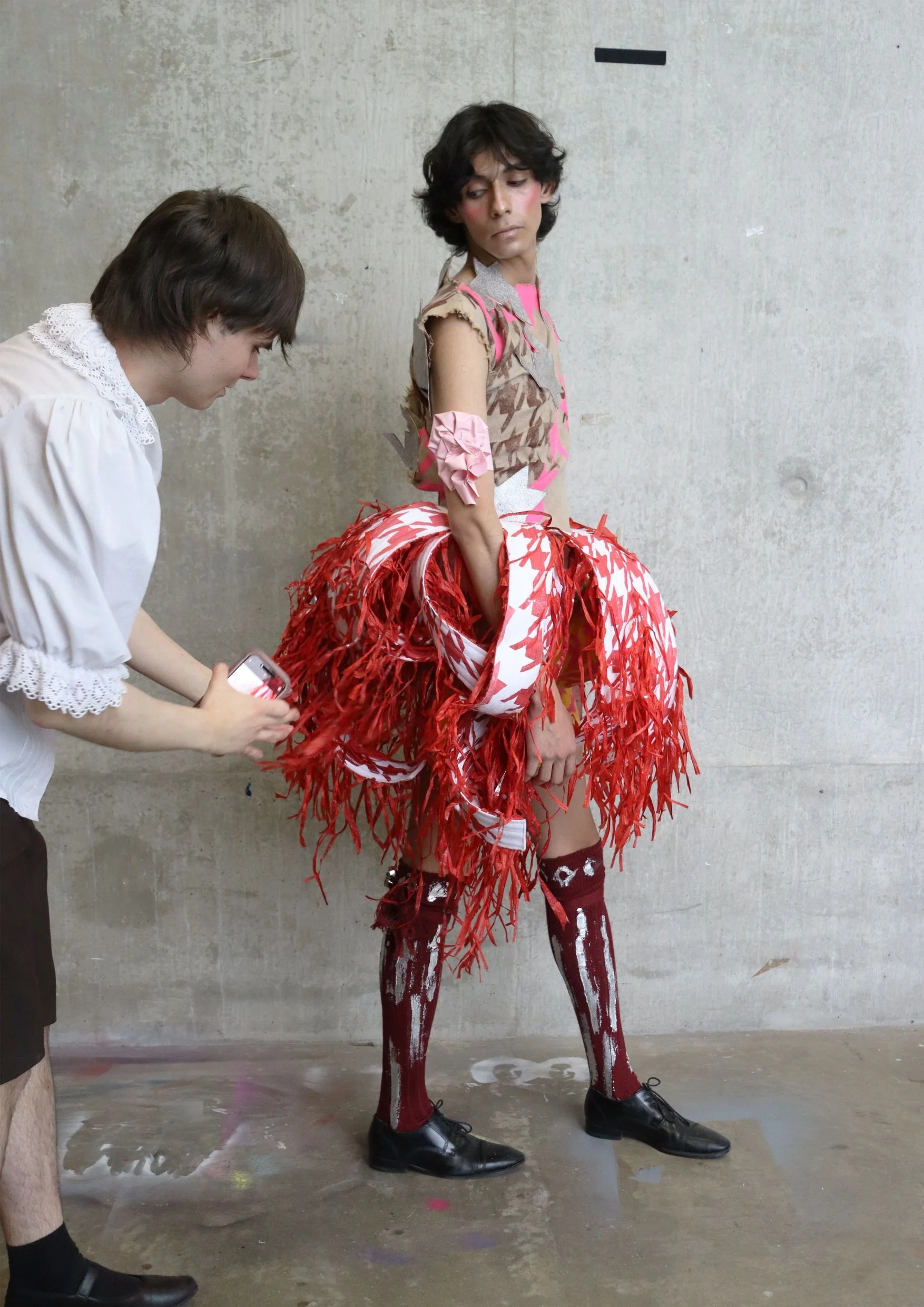 A person dressed in an avant-garde costume with a red feathered skirt, patterned top, and high socks, standing against a plain wall. Another person nearby appears to be adjusting or preparing the outfit, holding a phone. The setting seems to be a dressing room or backstage area.
