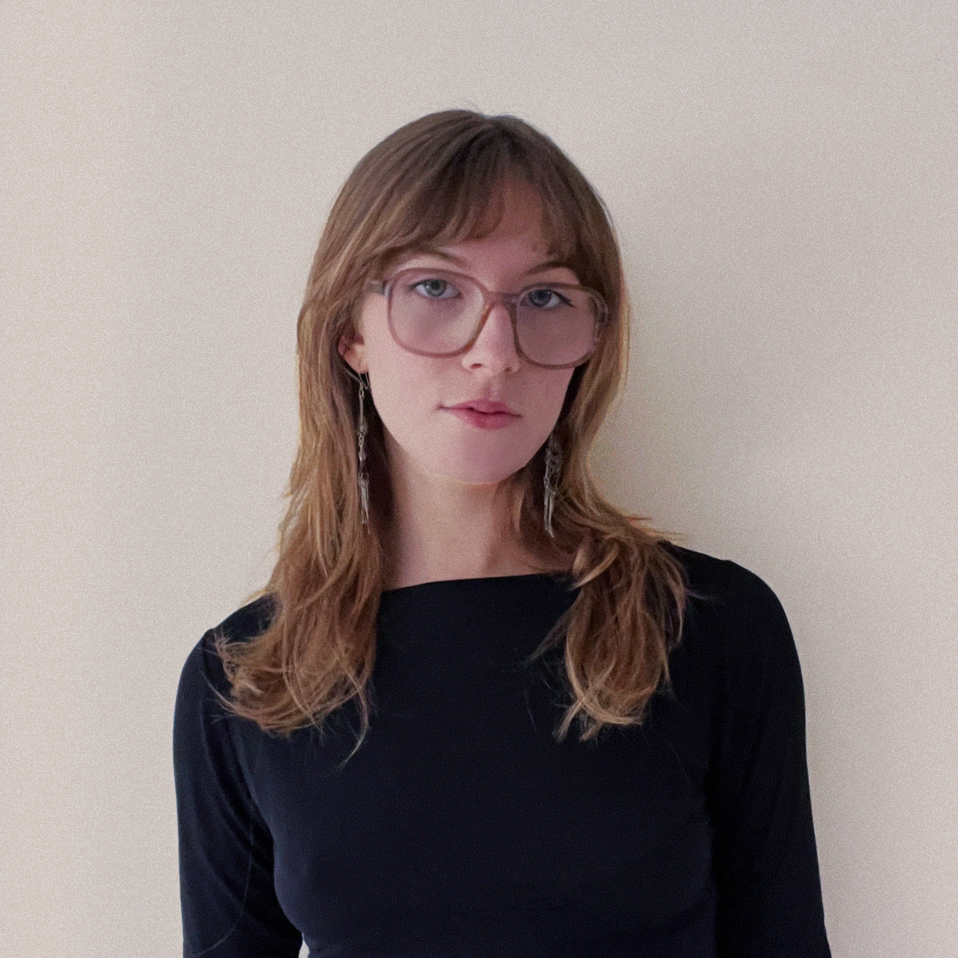 A woman with shoulder-length brown hair, wearing large pink glasses and earrings, is looking at the camera. She is dressed in a black top and is standing against a plain light-colored background.