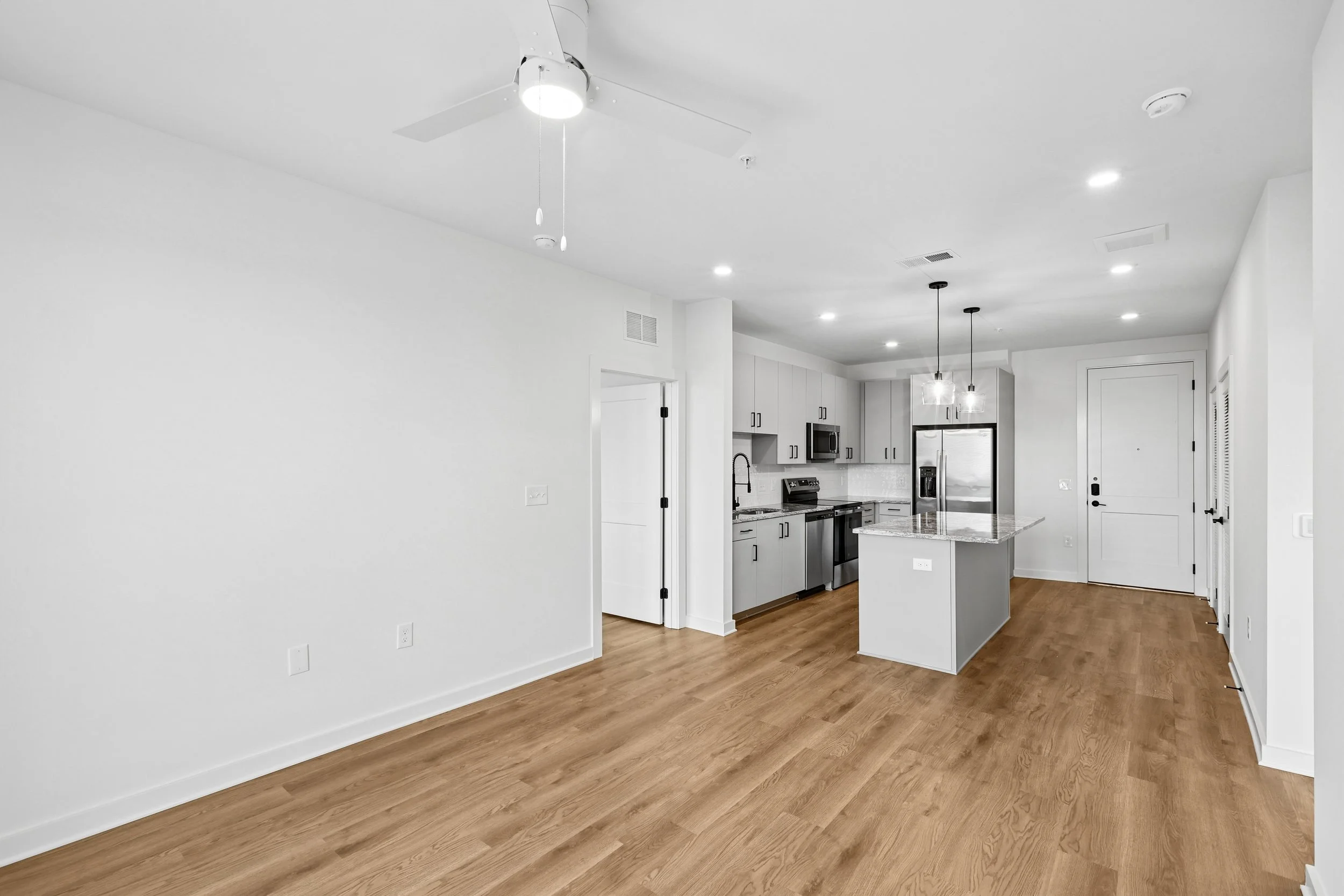 An open-concept kitchen with white cabinets, stainless steel appliances, and a granite countertop island. The space features light hardwood flooring, recessed lighting, and a ceiling fan.