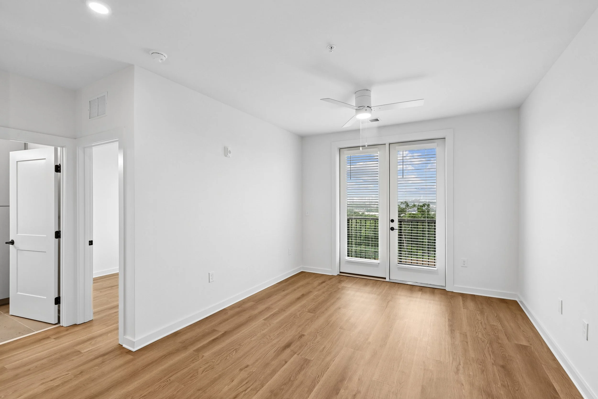 Empty room with white walls, wood flooring, a white ceiling fan, a glass door with blinds leading outside, and a window next to the door.