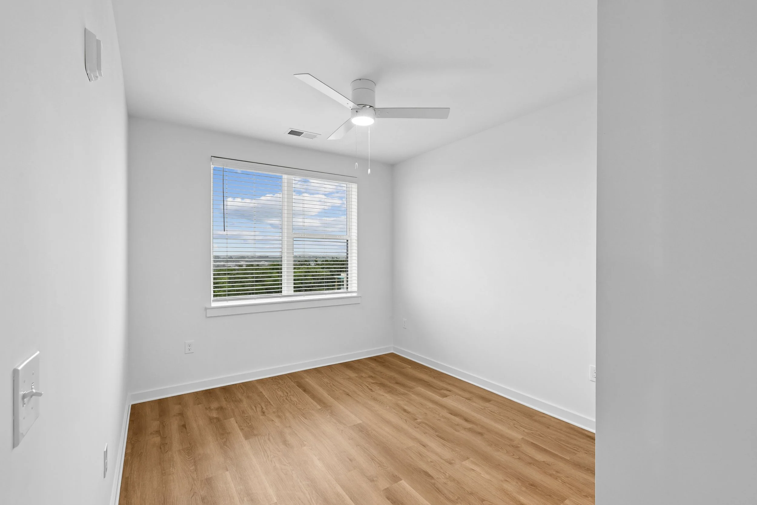 Empty room with white walls, hardwood floors, a window with blinds showing blue sky and clouds, and a white ceiling fan.