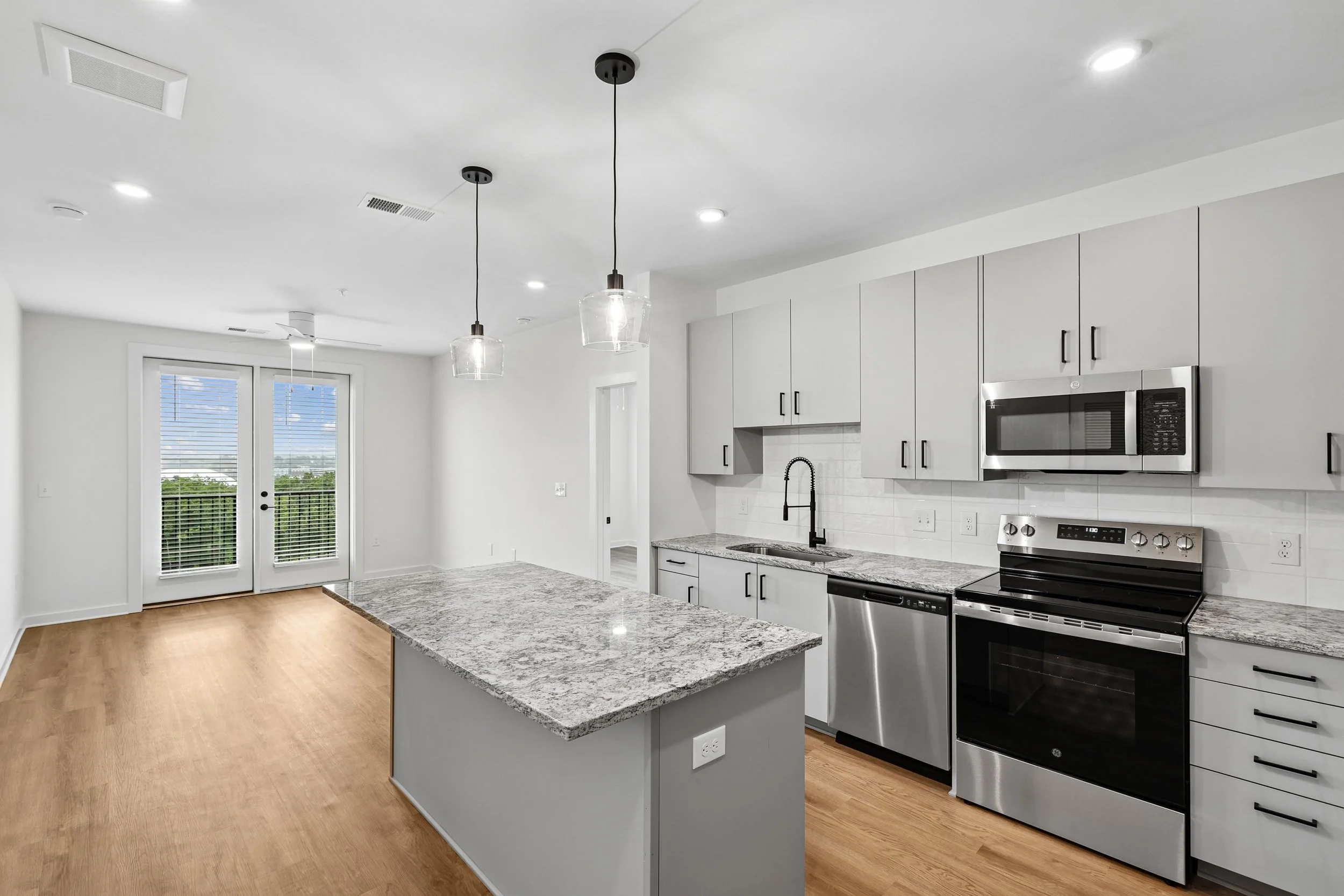 Modern kitchen with white cabinets, stainless steel appliances, granite countertops, pendant lights, and hardwood floors. A large window and glass door lead to an outdoor space.