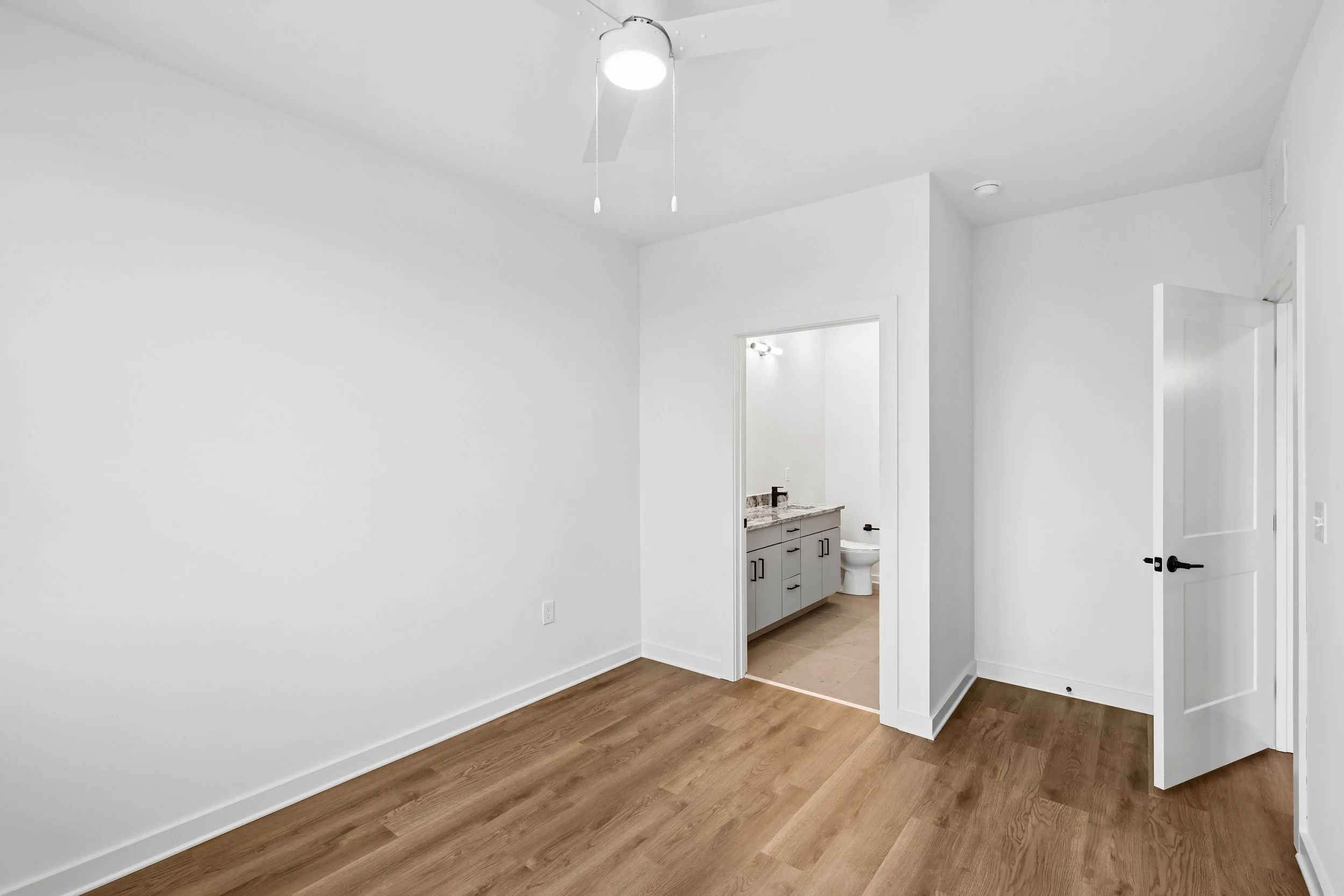 Empty room with white walls, light brown wood flooring, and a doorway leading to a bathroom with a vanity, granite countertop, and toilet; ceiling fan with light.