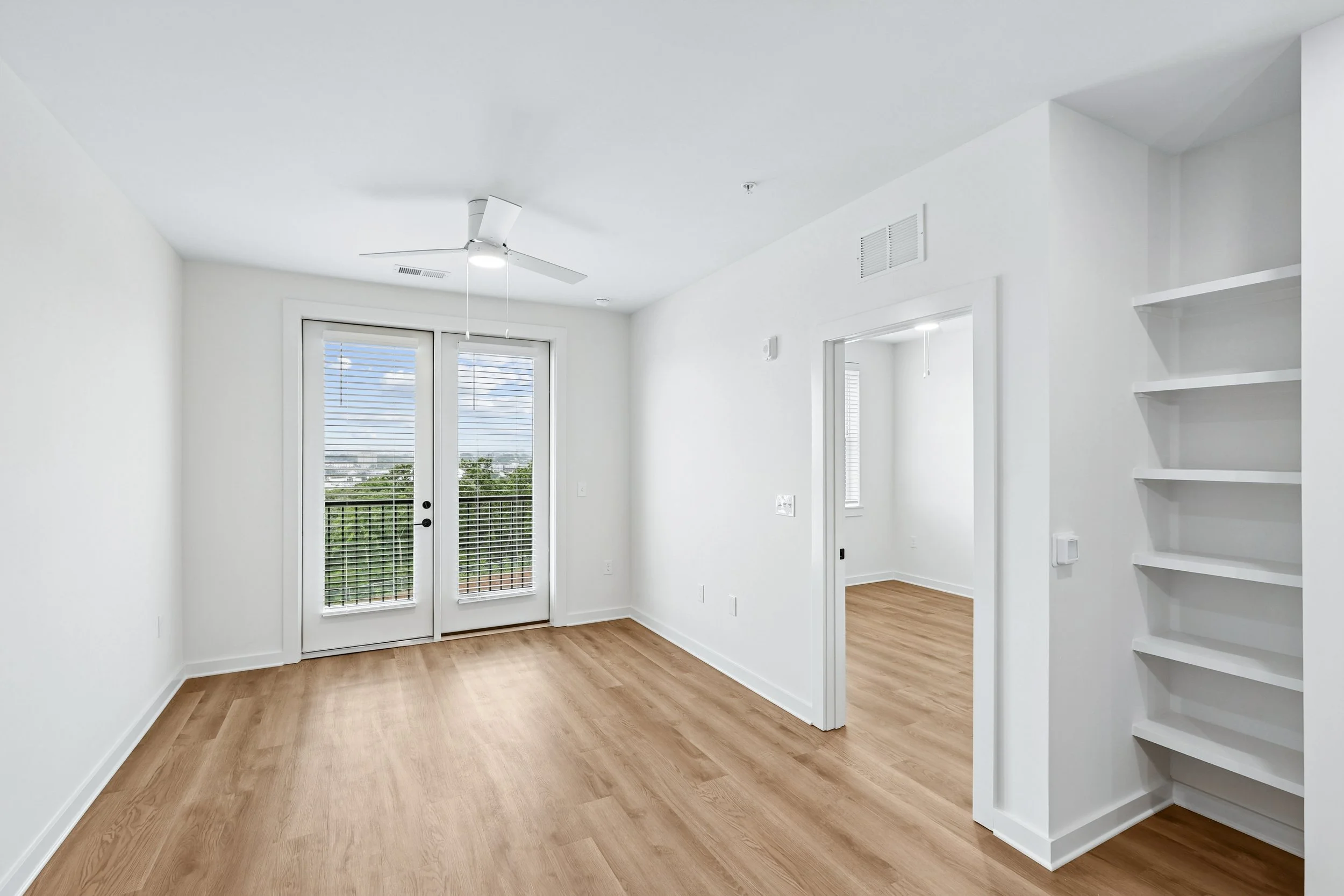 Interior of a modern, minimalistic living room with white walls, wood flooring, a ceiling fan, and glass doors leading outside. There are built-in shelves on the right side.