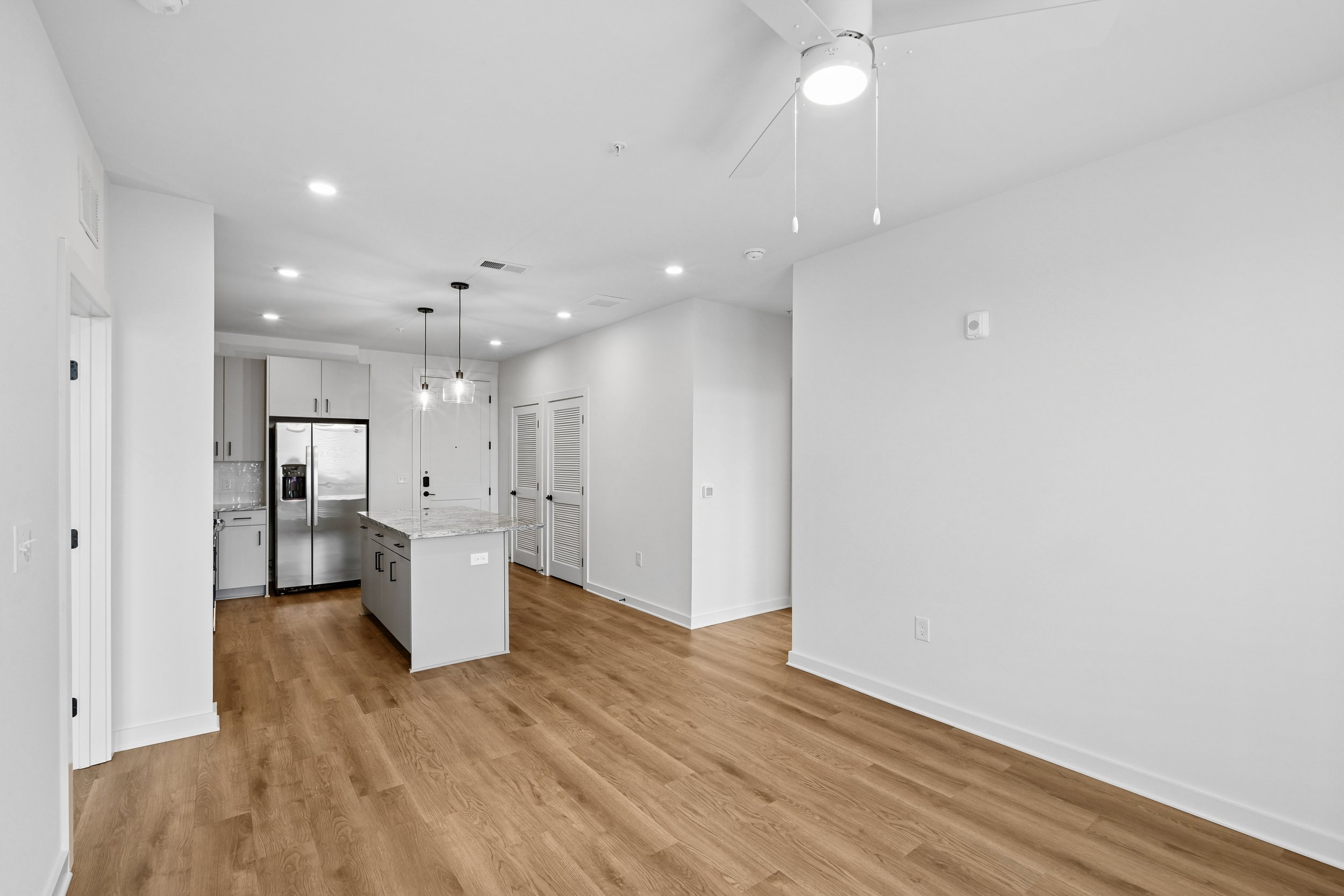 Empty modern living area with hardwood floors, white walls, recessed ceiling lights, ceiling fan, and a kitchen with white cabinets, stainless steel refrigerator, and a breakfast island.