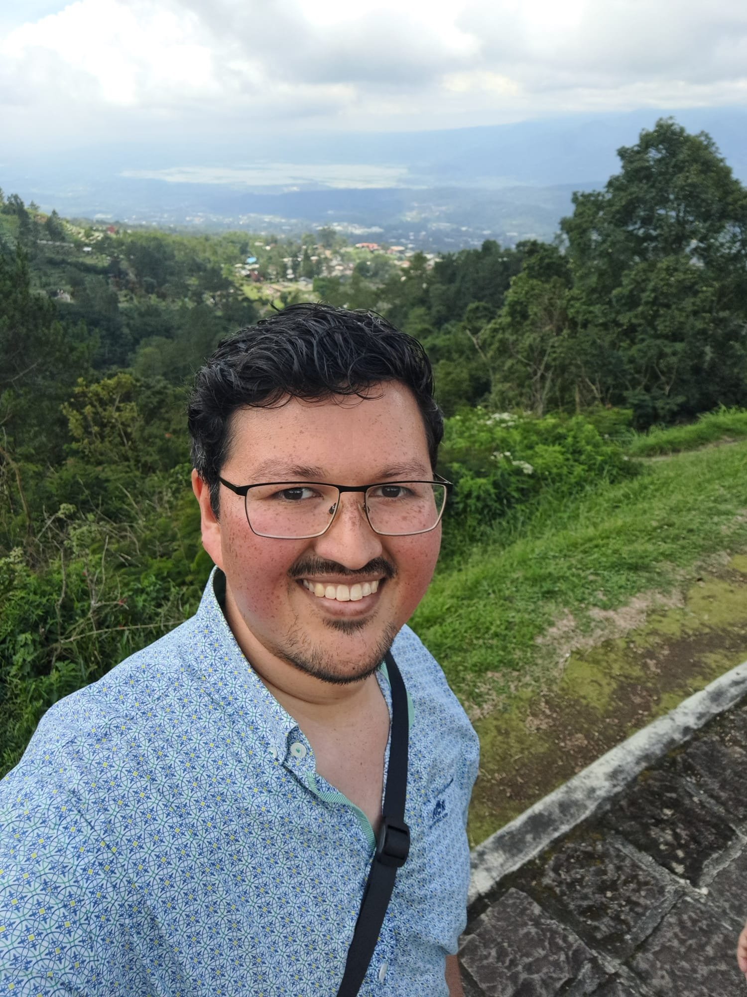Man met donker haar en bril, die een een grote lach op zijn gezicht heeft en een blauwe shirt draagt, staat buiten met een groen landschap en bergen op de achtergrond. Cliënt van Spectrumworks: Kevin. 