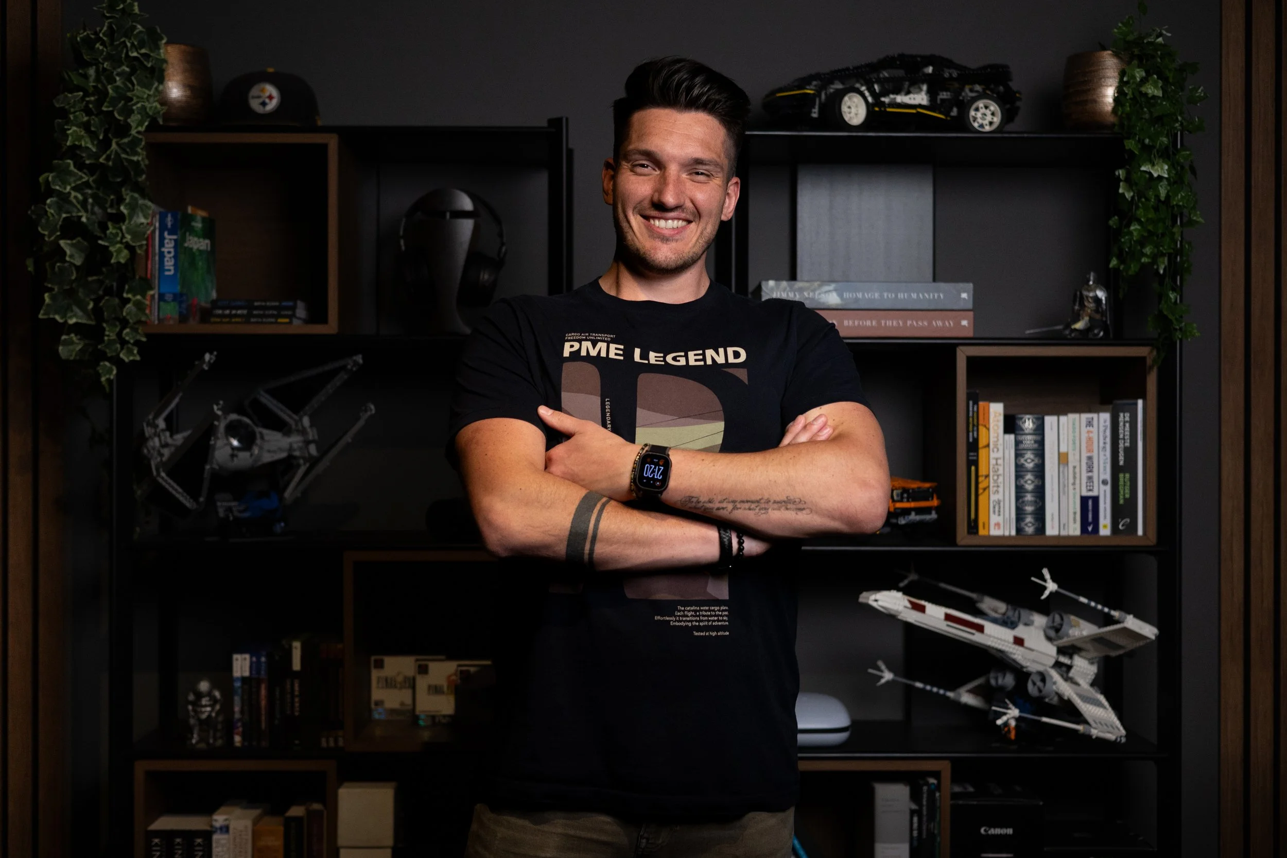 Vrolijke jonge man met donker haar, zwart T-shirt en smartwatch, staand met armen gekruist voor een zwarte boekenkast vol boeken, LEGO-model en planten. Betreft de eigenaar van Spectrumworks: Raymon Boel