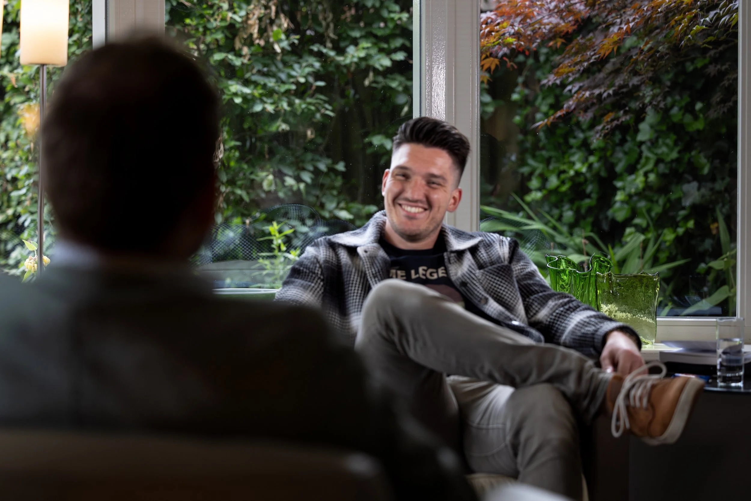 Man die in gesprek is, eigenaar van Spectrumworks met zijn cliënt Marcel. Zij zitten samen in een woonkamer, waarbij op de achtergrond de ramen en de tuin te zien zijn.