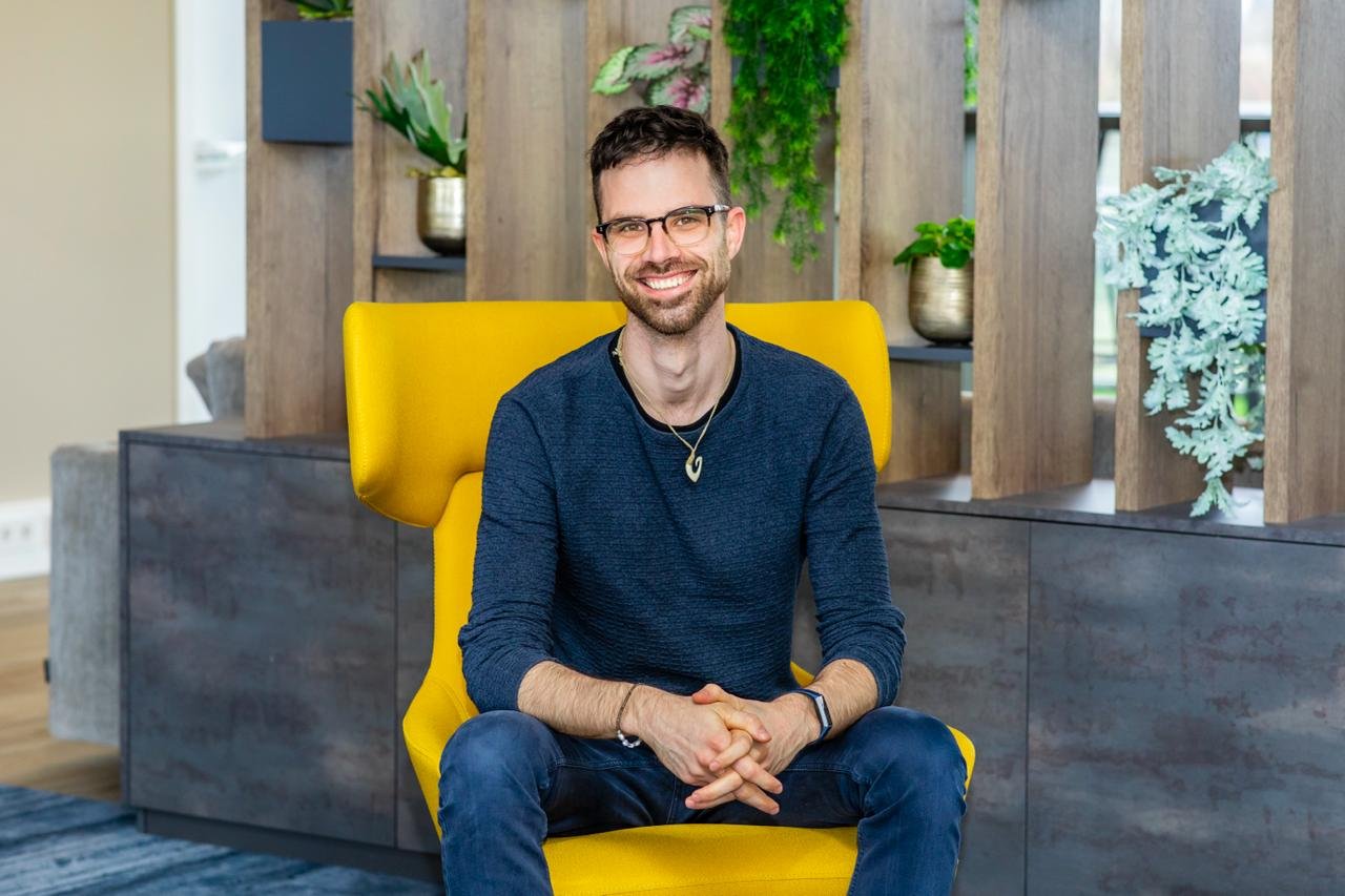 Man zit op een gele stoel, lachend, in een binnenruimte met planten op de achtergrond. Een cliënt van Spectrumworks: Rhys. 
