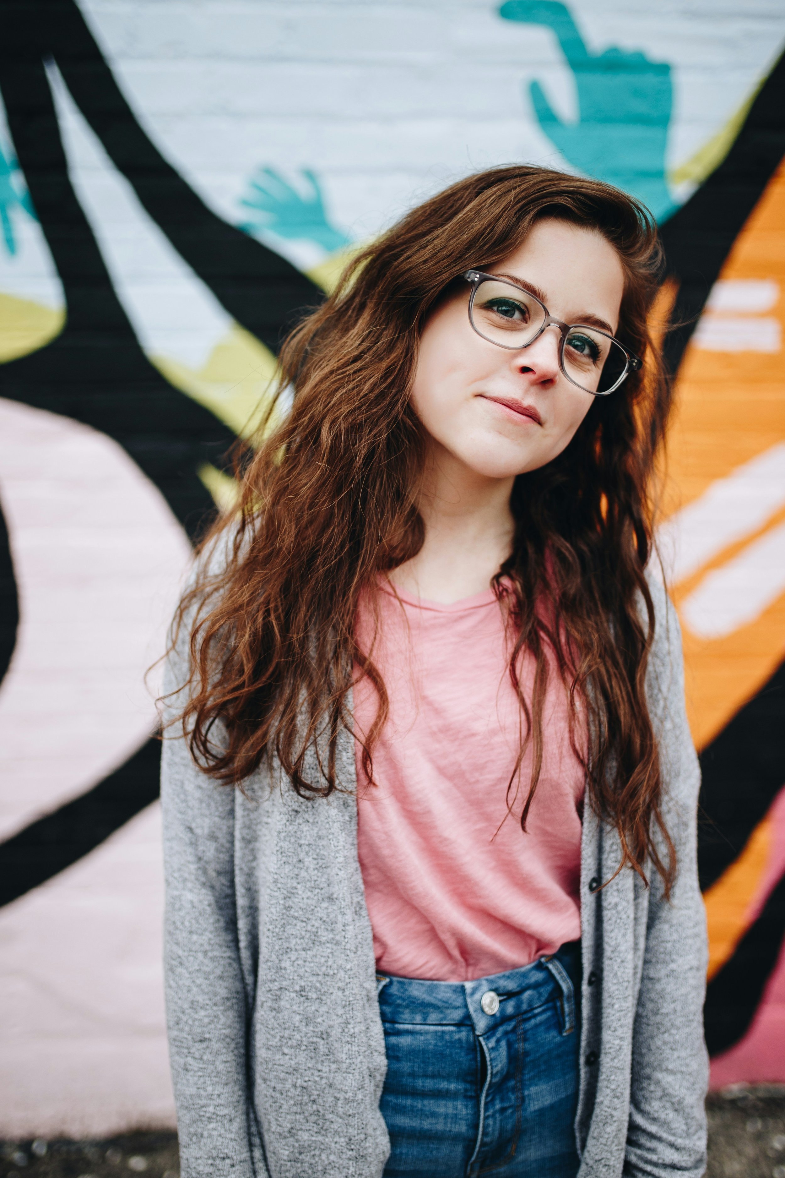 Portret van een jonge vrouw met lang bruin haar, bril, roze T-shirt en grijze jas voor een kleurrijke muurschildering. Hierbij duidt de eigenaar op een tevreden cliënt; iemand die tevreden is met de begeleiding van Spectrumworks.