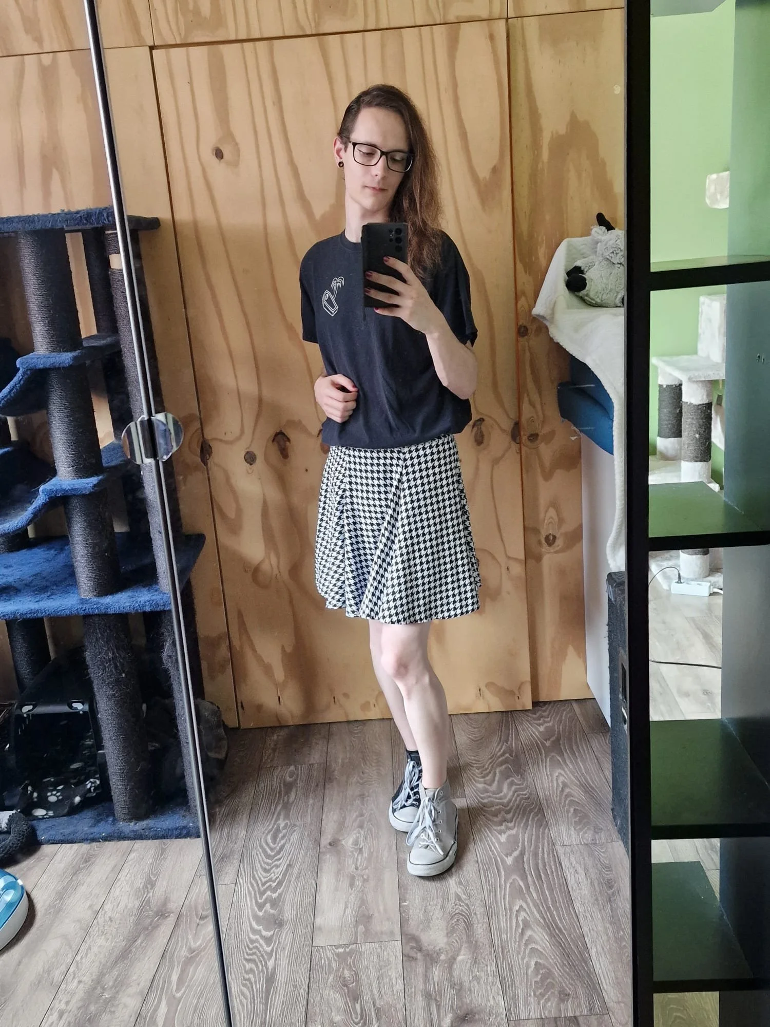 Een vrouw met lang haar, bril en piercings, gekleed in een zwart T-shirt en een zwart-wit geruite rok, neemt een selfie in een spiegel met een smartphone. Ze staat in een kamer met houten panelen op de muur, een kattenkrabpaal links en een kast met een doek en een knuffel rechts.
