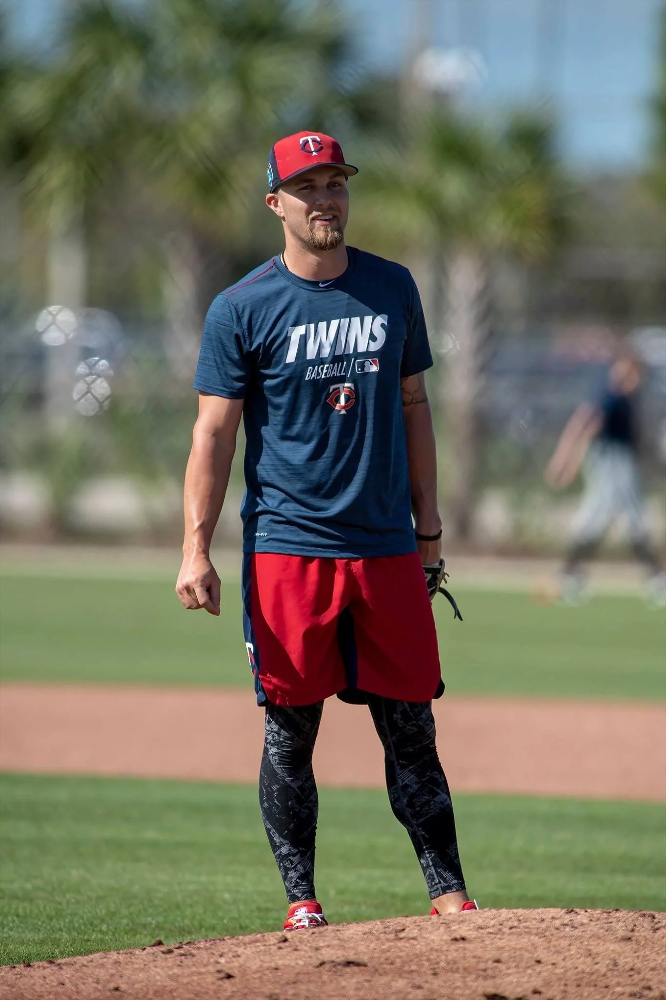 A man standing on a baseball field wearing a Twins baseball shirt, red and black shorts, and leggings, with a baseball glove on his left hand and a baseball cap.