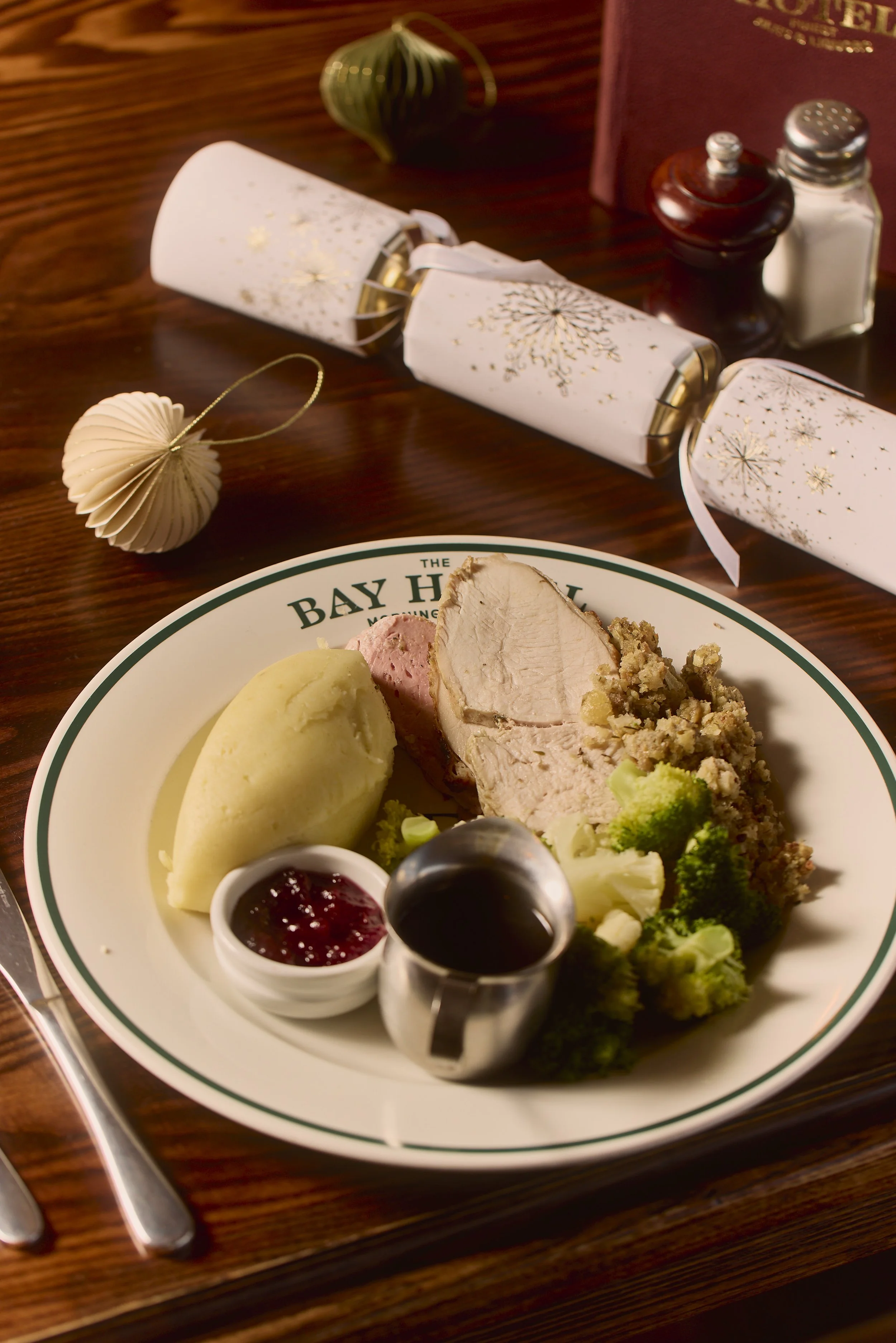 A plate of food with mashed potatoes, slices of turkey, broccoli, and cranberry sauce. In the background, a holiday-themed turndown service with decorative gold snowflakes on white paper scrolls, a paper bauble, and salt and pepper shakers on a wooden table.
