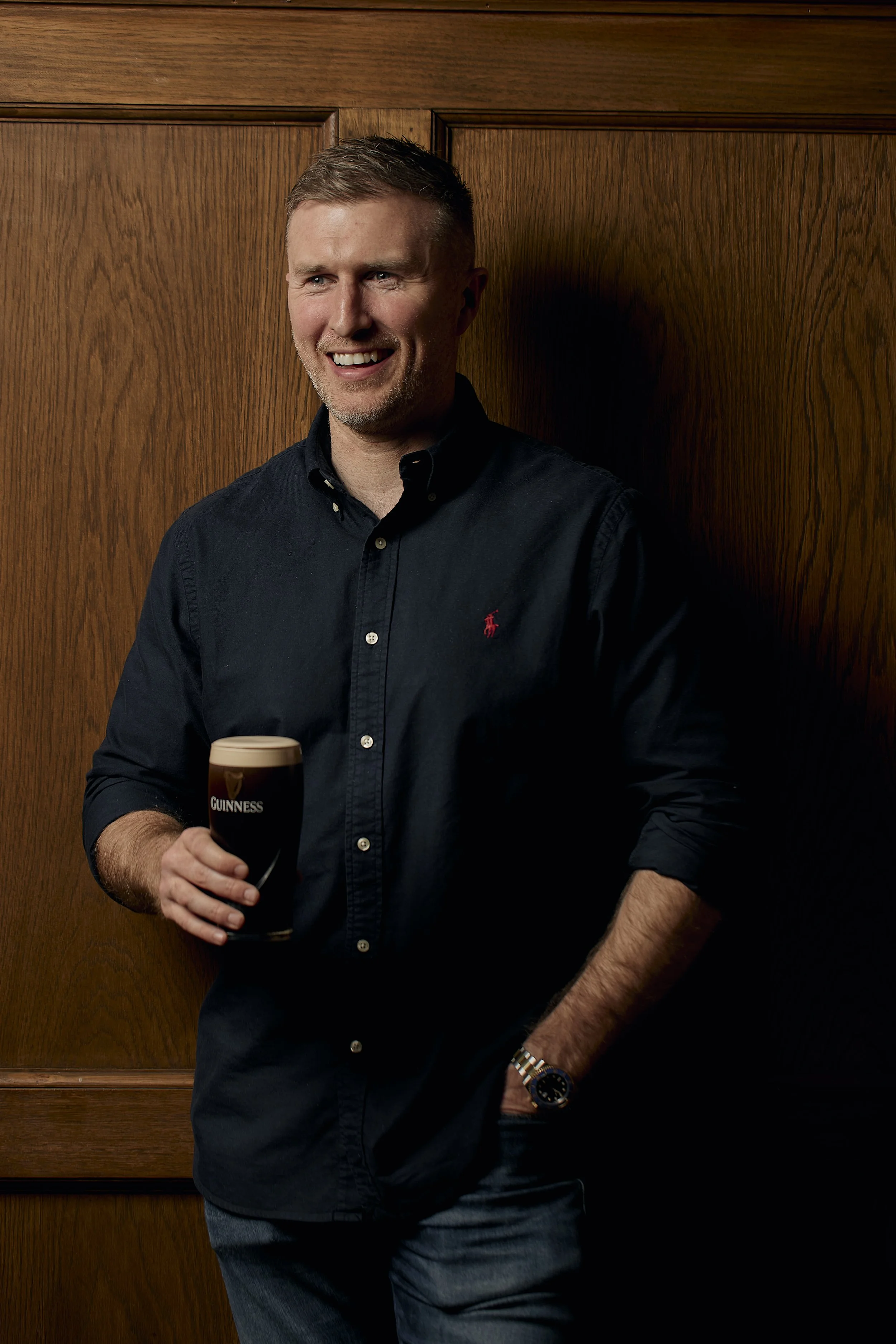 A man with short hair, light skin, and a beard is holding a pint of Guinness beer while smiling. He is standing in front of a wooden wall, wearing a navy blue shirt and a wristwatch.