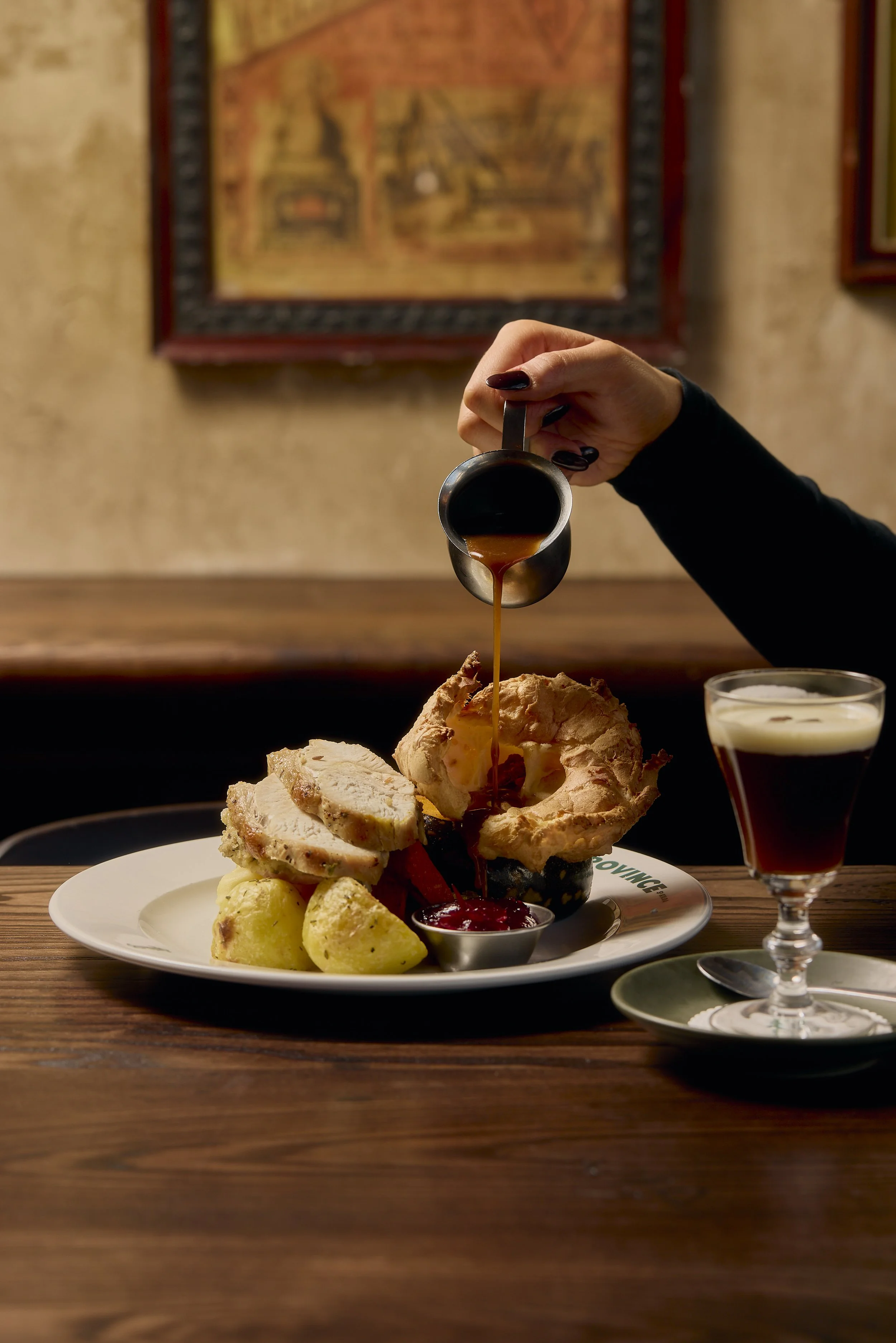 A person pouring gravy over a large portion of carved turkey at a Thanksgiving feast, with mashed potatoes, cranberry sauce, and a glass of dark beverage on a wooden table.