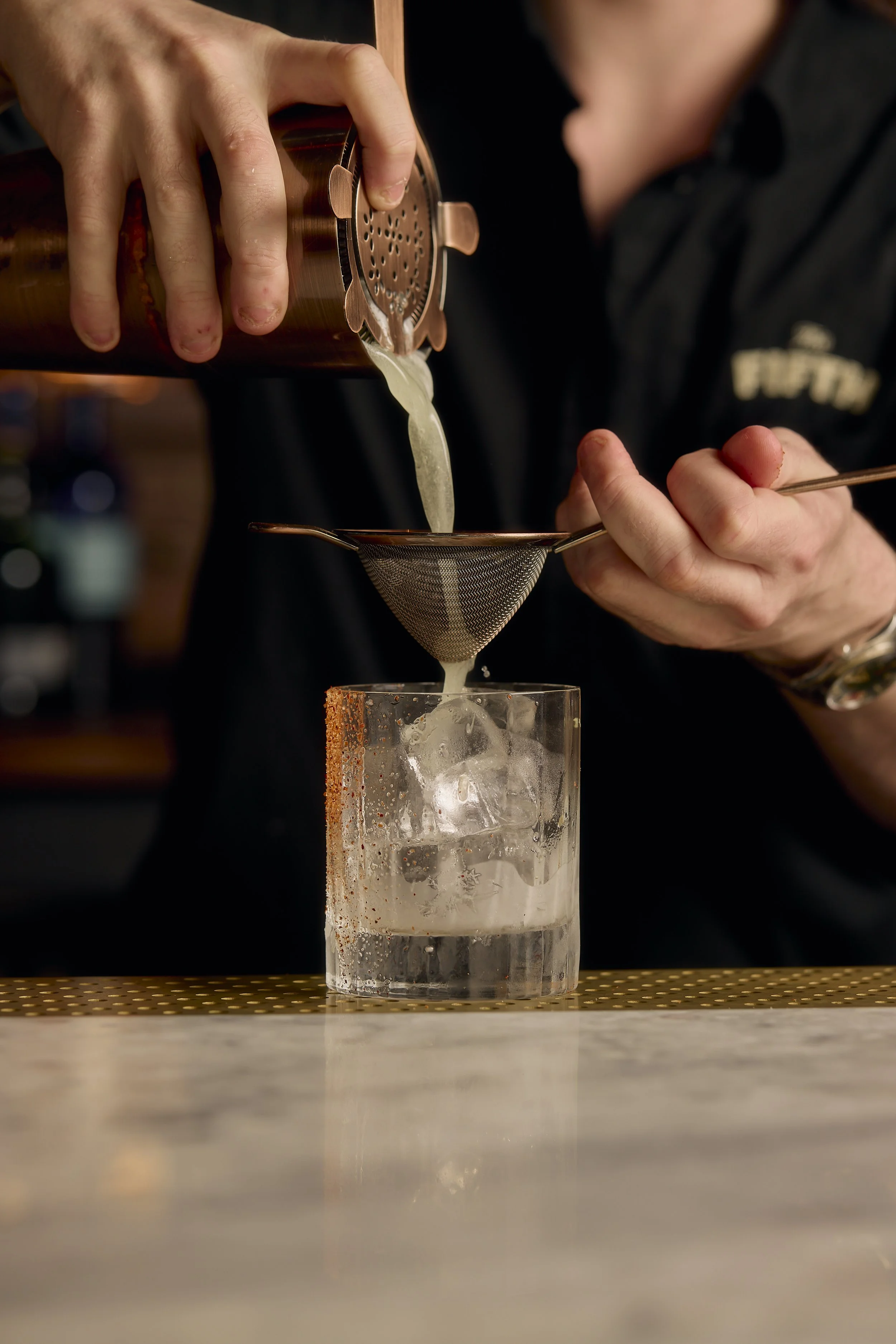 Bartender pouring liquid through a cocktail strainer into a glass with ice cubes.