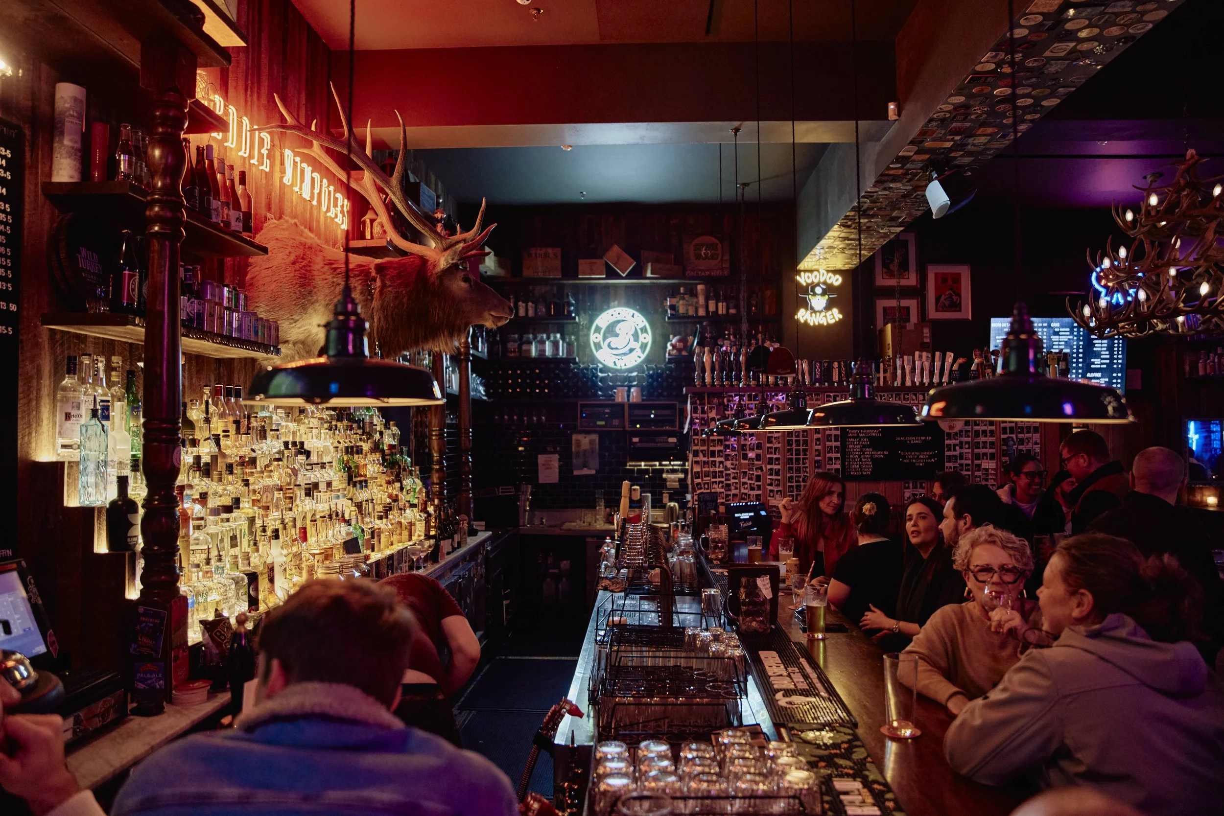 A busy bar with a well-lit counter displaying numerous bottles of liquor, a mounted deer head with antlers, and a neon sign in the background. Several patrons are sitting and standing, engaged in conversations, with various decorations including a chalkboard menu and framed pictures on the walls.