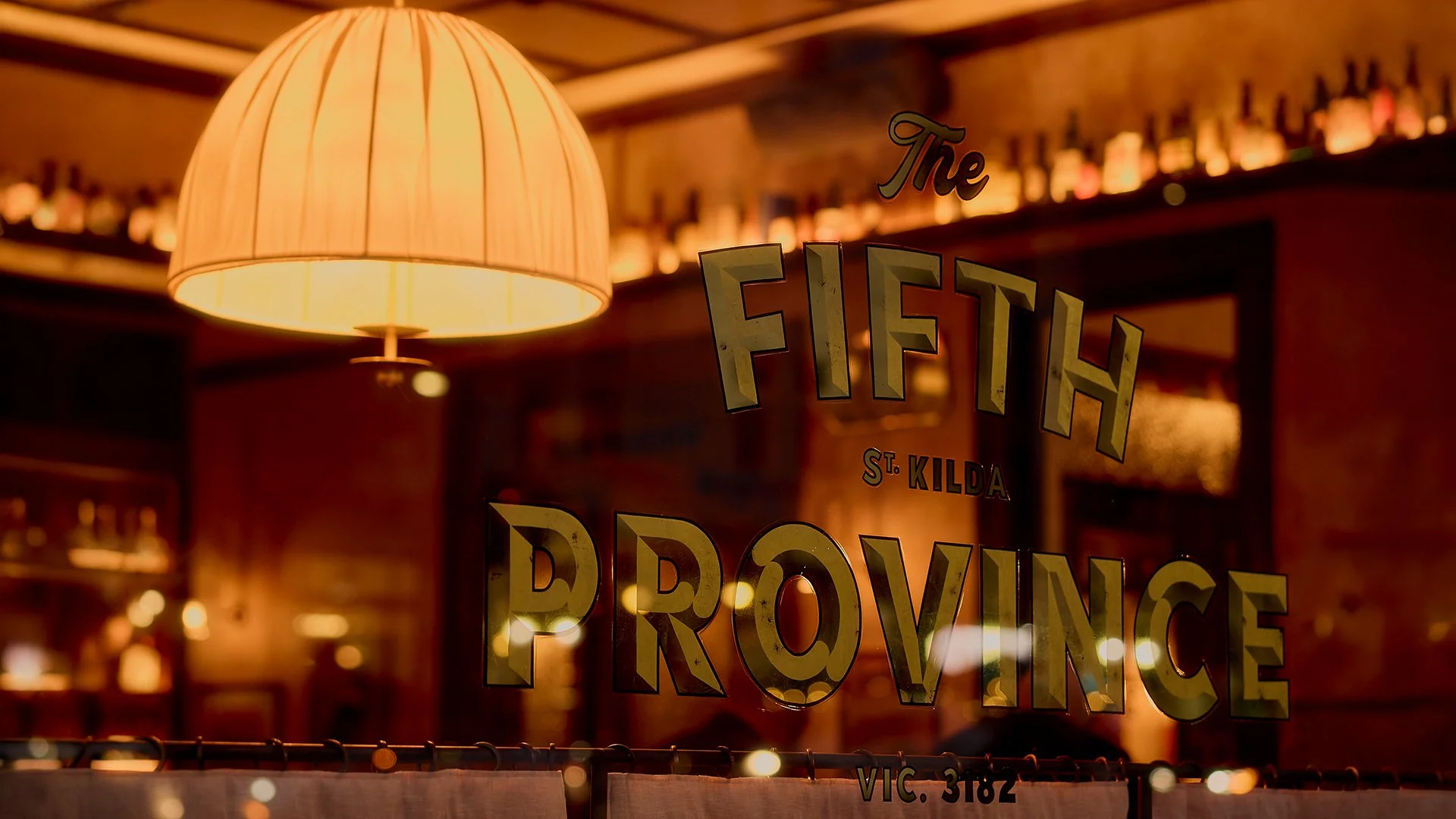 A warm-lit bar with a hanging beige lampshade. There is a glass window reflecting the bar's name, 'The Fift H', with part of the text visible including 'St. Kilda' and 'Provence', along with some bottles on a shelf in the background.
