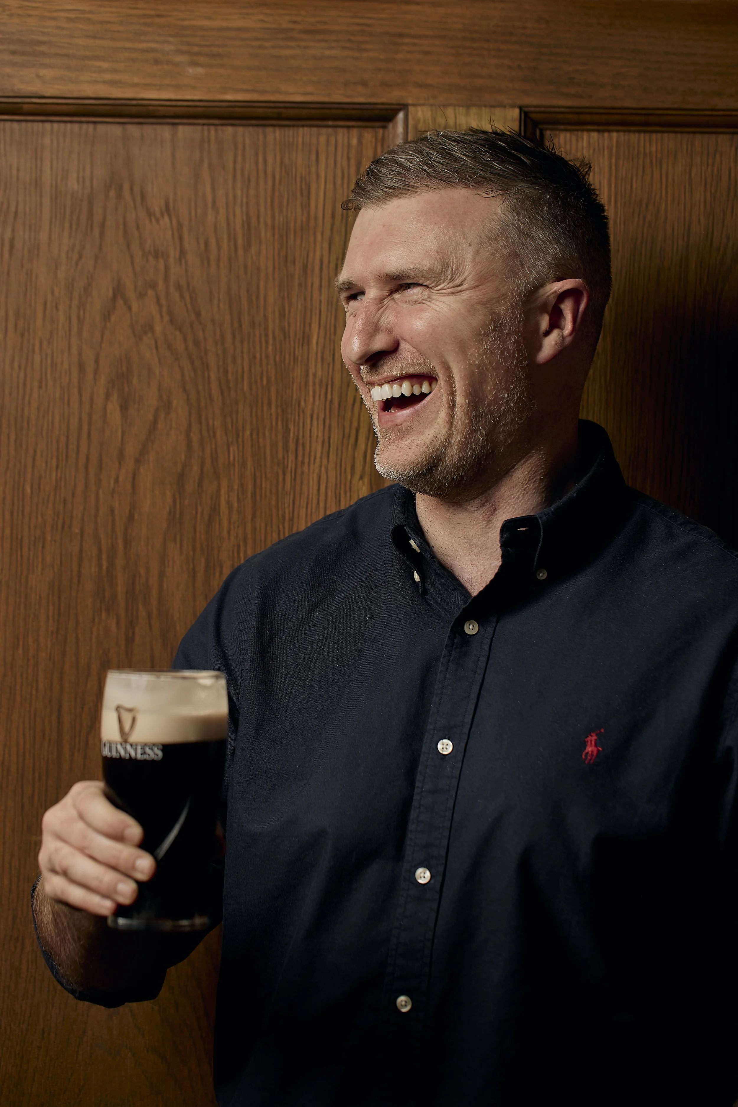 A smiling man with short hair and a beard, wearing a dark button-up shirt, holding a pint of Guinness beer, standing in front of wooden paneling.