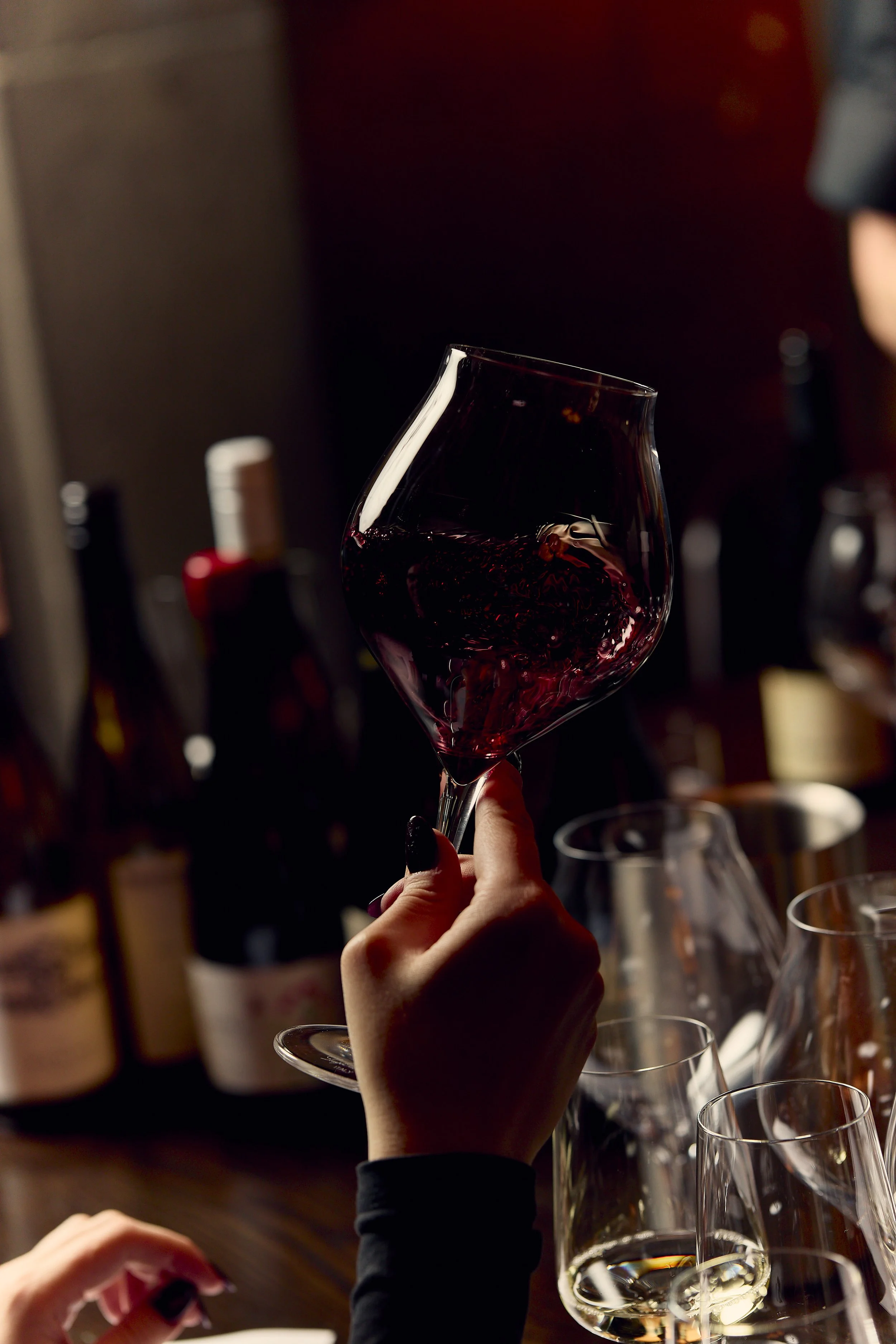 A person holding a glass of red wine at a social gathering, with several wine bottles and glasses on a table in the background.