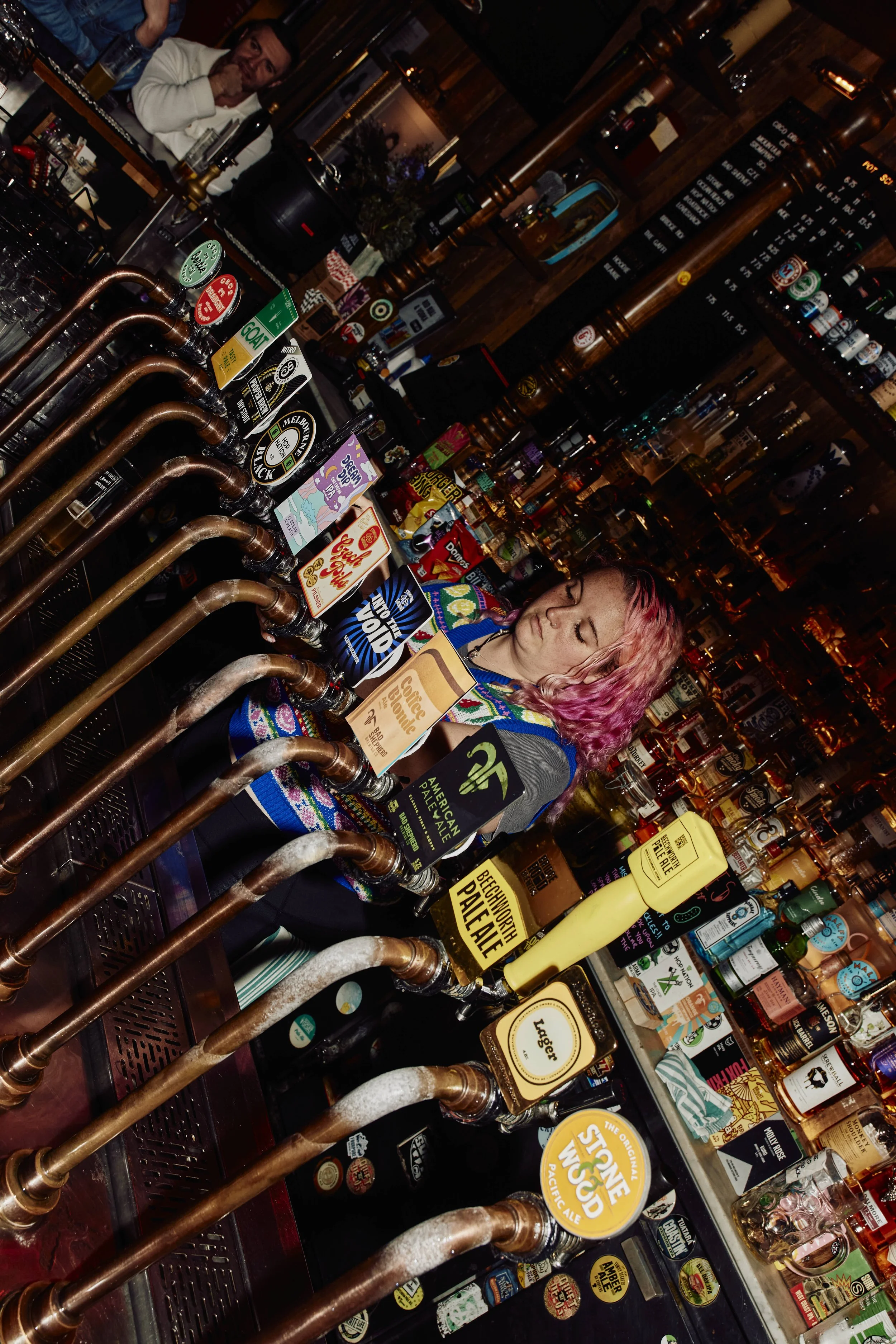 A bar with a woman with pink hair, wearing a colorful sweater, standing behind the counter. The bar has various taps for different beers and numerous bottles on shelves behind. A man with a beard and glasses can be seen in the background.