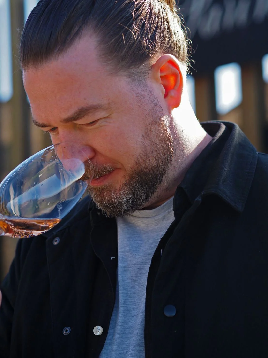 A man with a beard and long hair tied back, smelling a glass of rosé wine outdoors.