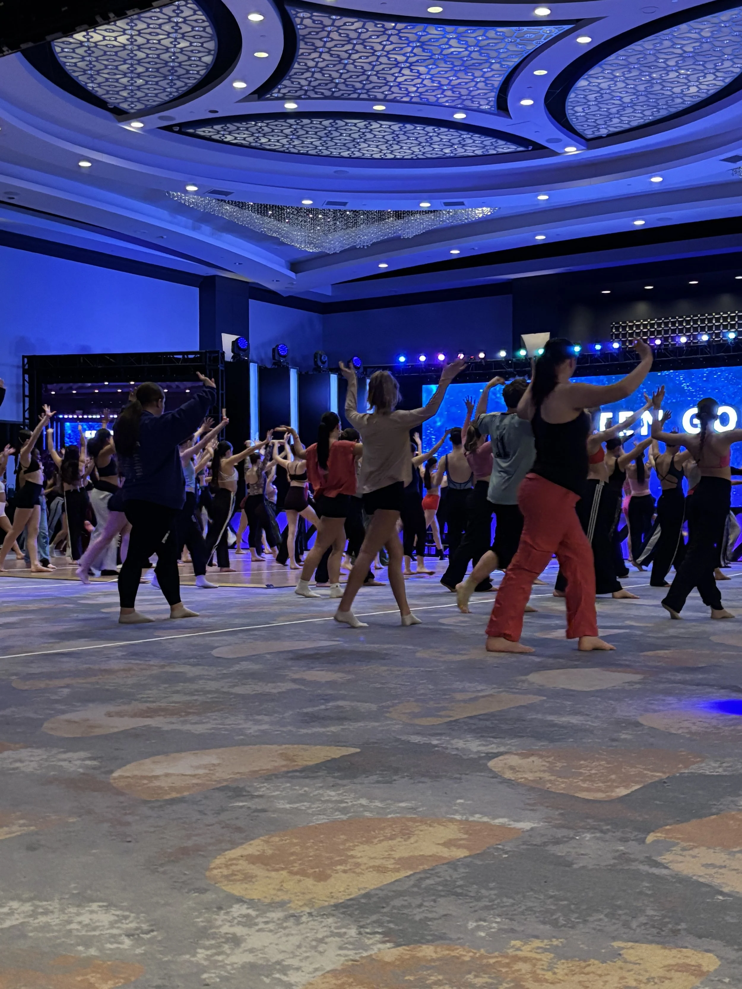 Group of people practicing dance or aerobics in a large, well-lit event hall with a decorative ceiling and a digital backdrop.